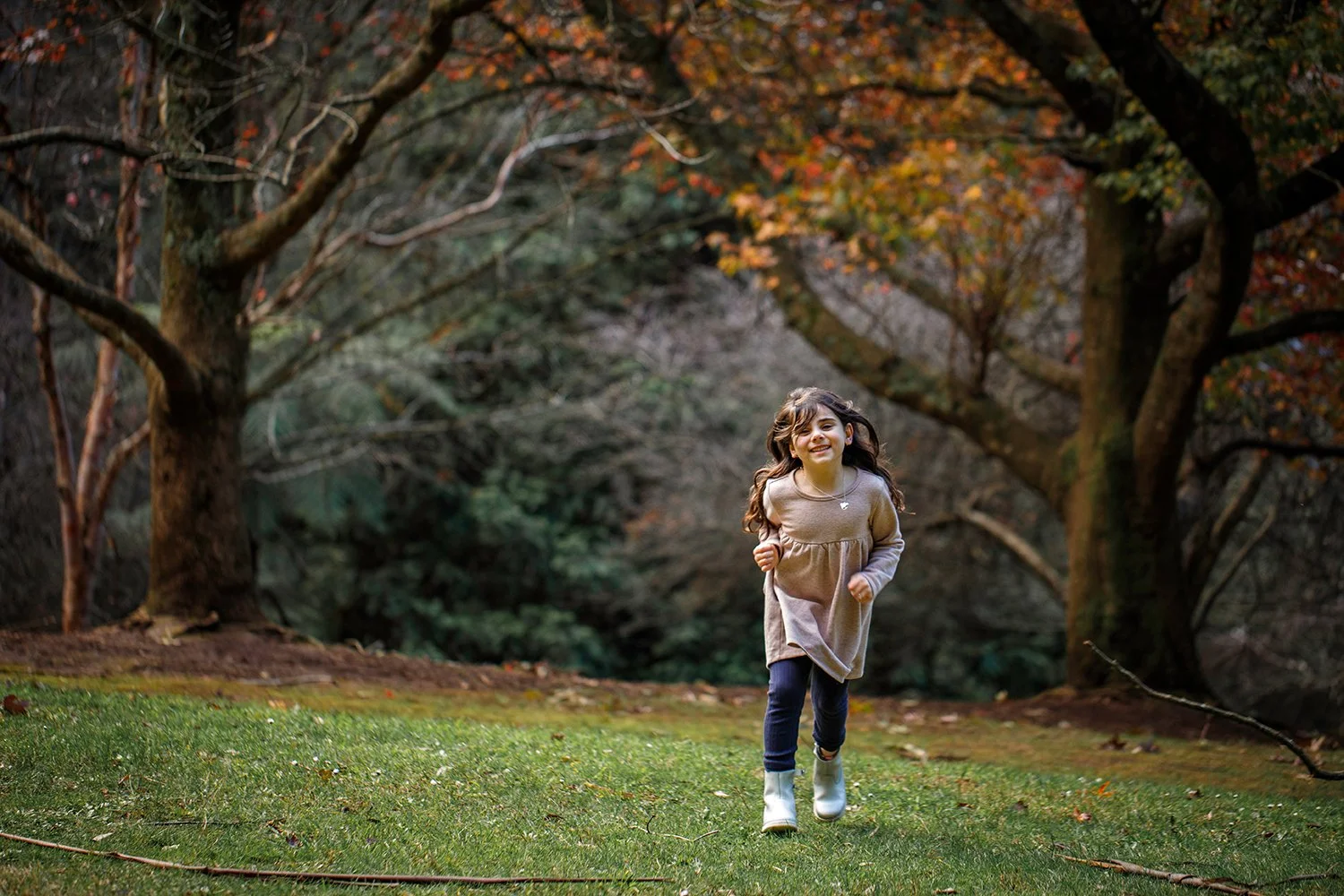 A young girl running on a grassy area in a park with trees that have autumn-colored leaves in the background.