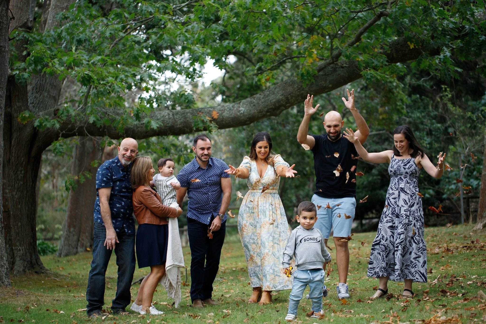 A group of seven people, three adults, and four children, are outdoors in a park playing with fallen leaves under a large tree during daytime.
