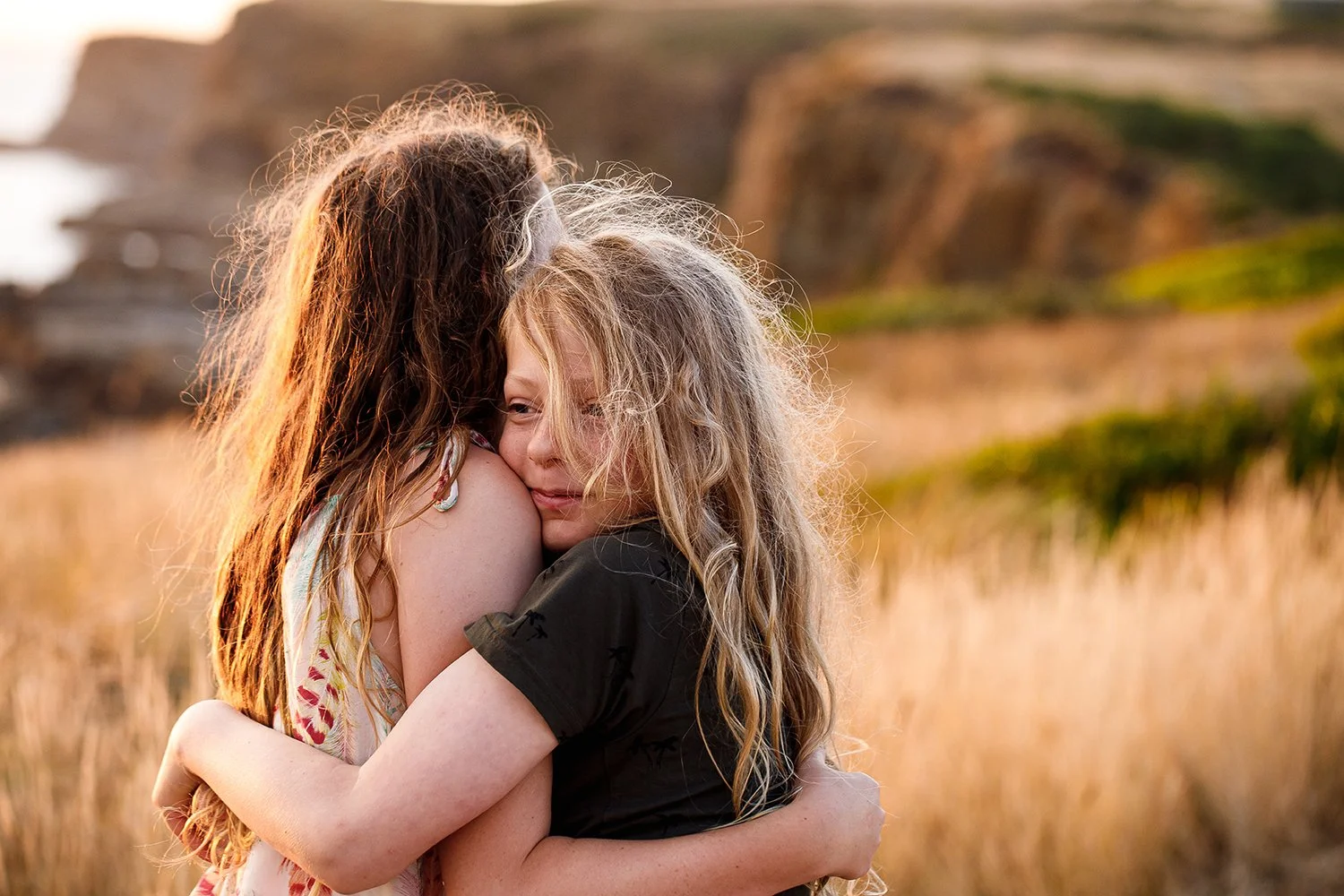 Two young girls hugging each other in a field at sunset, with soft golden light and blurred cliffs in the background.