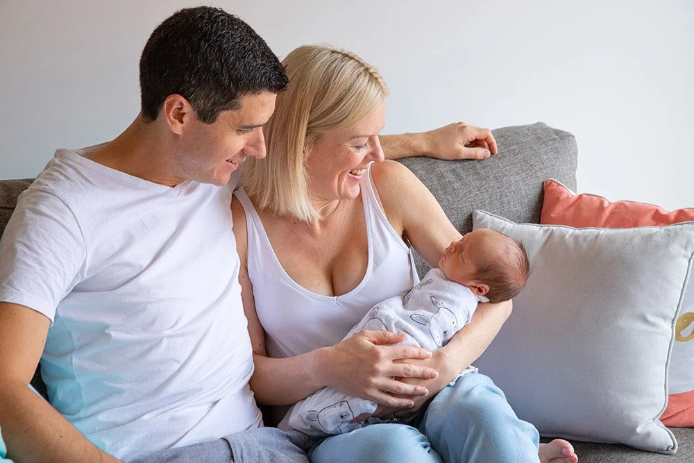 A smiling woman with blonde hair in a white tank top holding a newborn baby, sitting on a gray couch with a man in a white T-shirt, in a cozy living room.