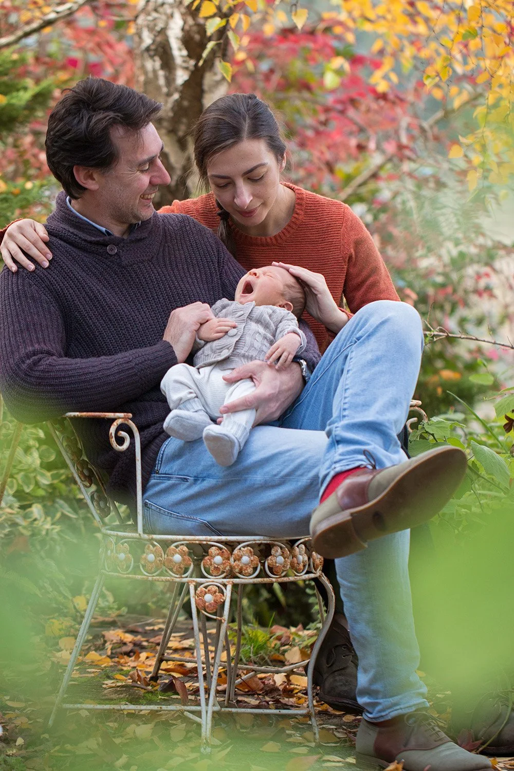A couple sitting on a decorative bench outdoors in autumn, holding and playing with a yawning baby surrounded by colorful fall foliage.
