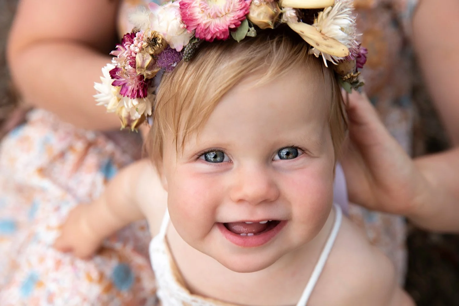 A young child with blonde hair and blue eyes smiling, wearing a floral crown and a sleeveless dress, outdoors.