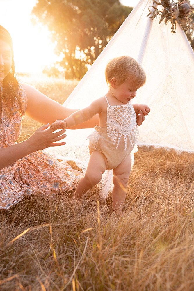 A baby learning to walk outdoors at sunset, holding hands with a woman in a field with tall grass. There is a white teepee decorated with flowers in the background.