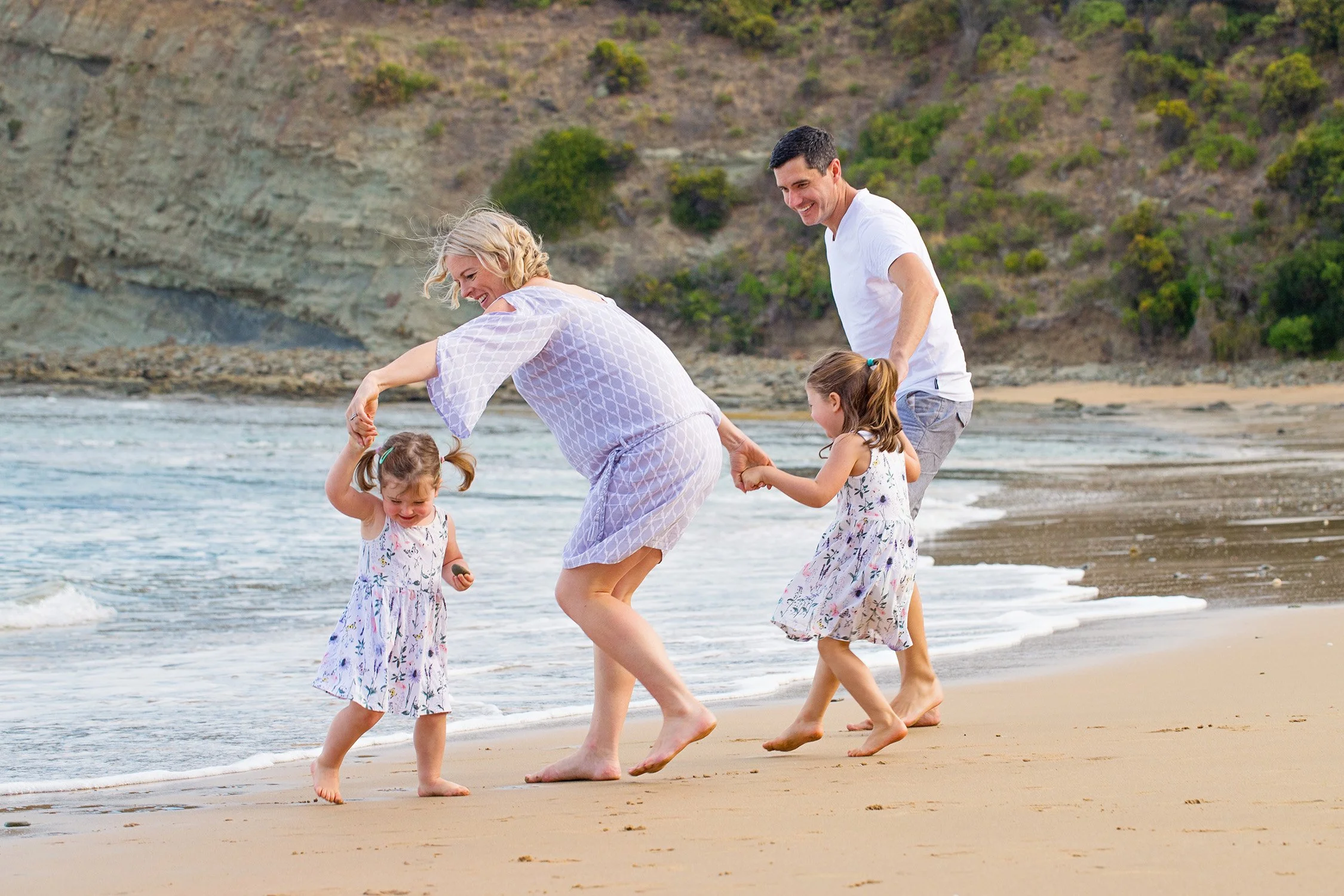 A happy family of four enjoying time at the beach, with two young girls playing and their parents holding hands near the shoreline, with a backdrop of hills and greenery.