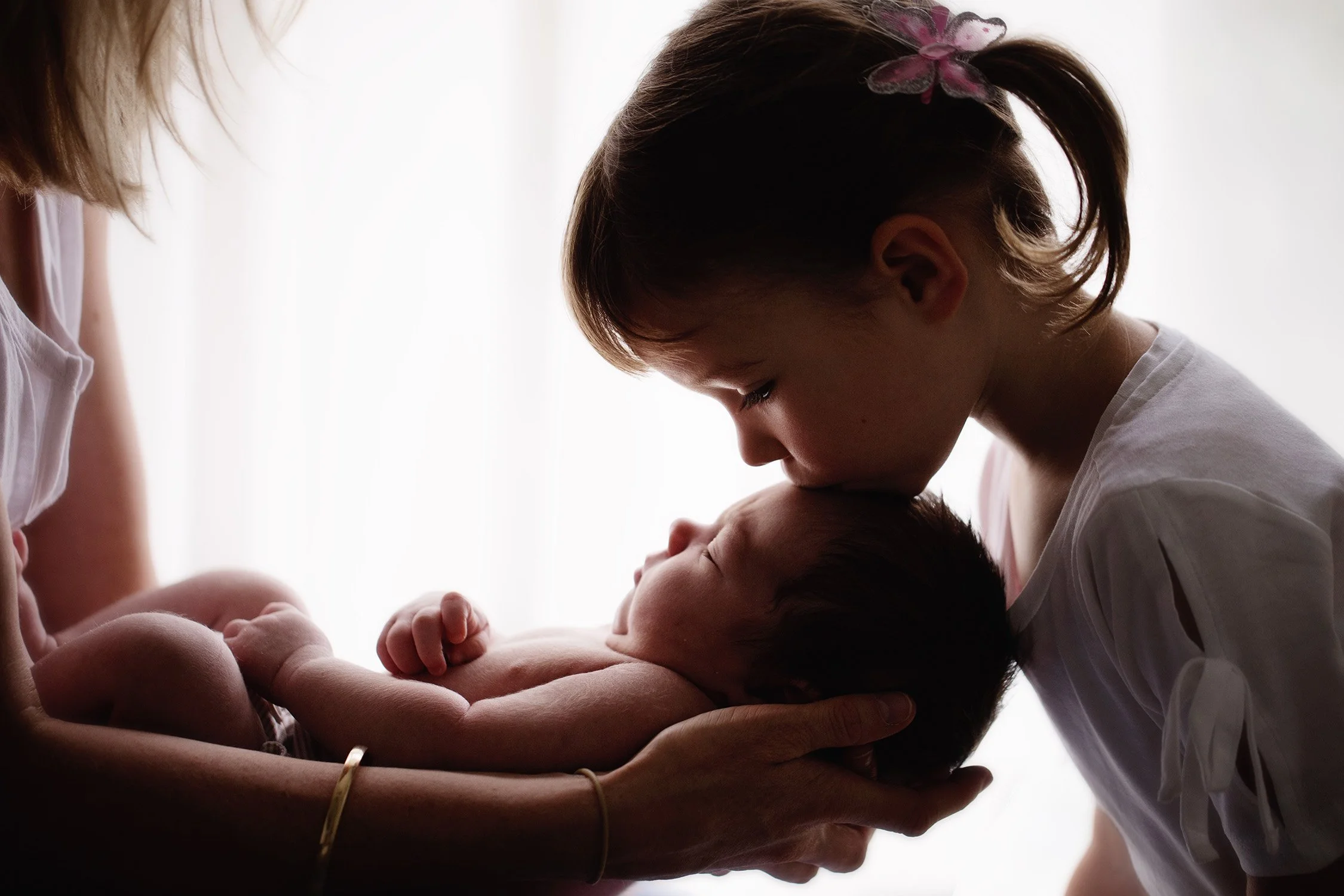 A young girl gently kissing a newborn baby on the forehead, with an adult holding the baby.