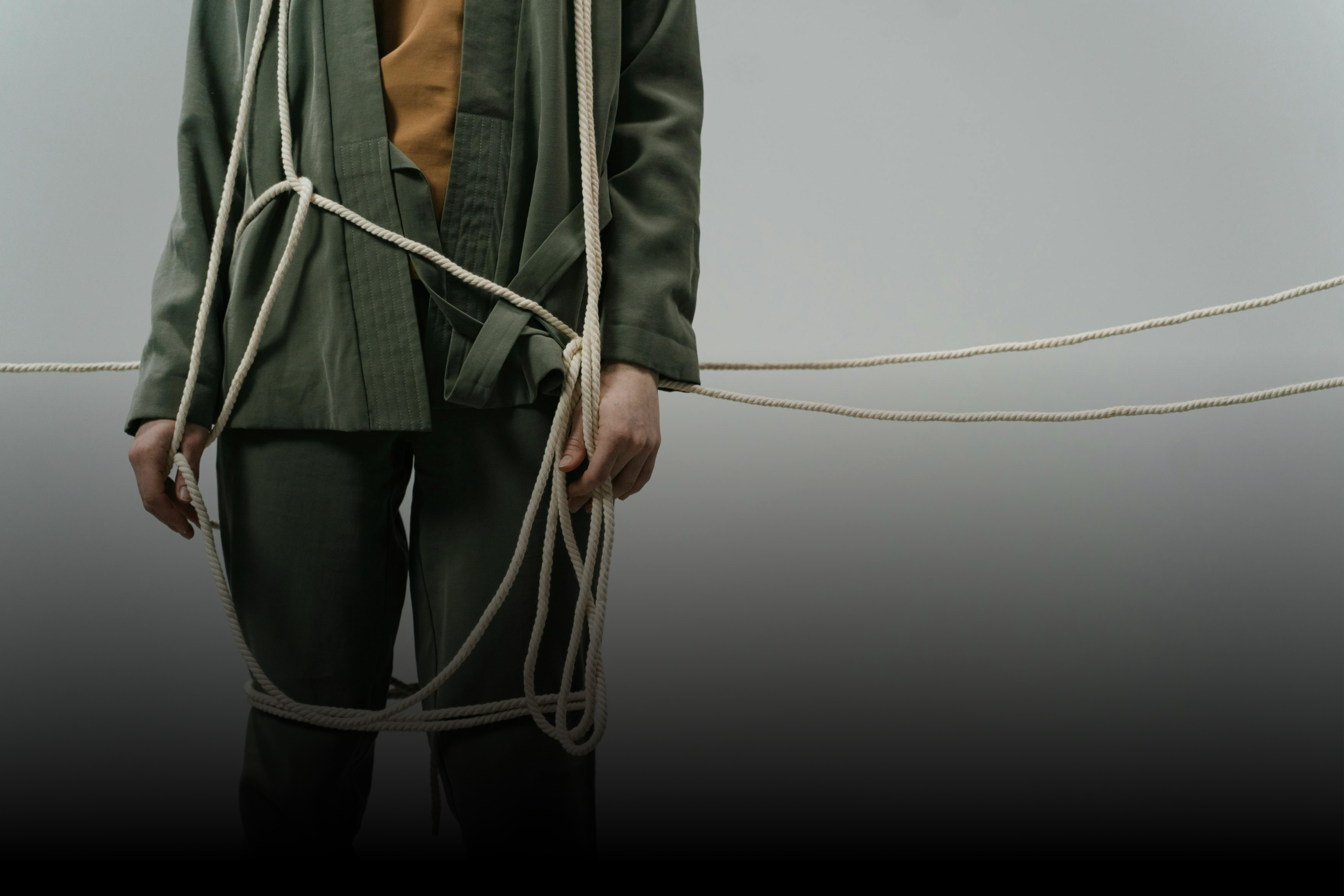 Person wearing green clothing, holding a thick rope, with more ropes stretching horizontally across a plain background.