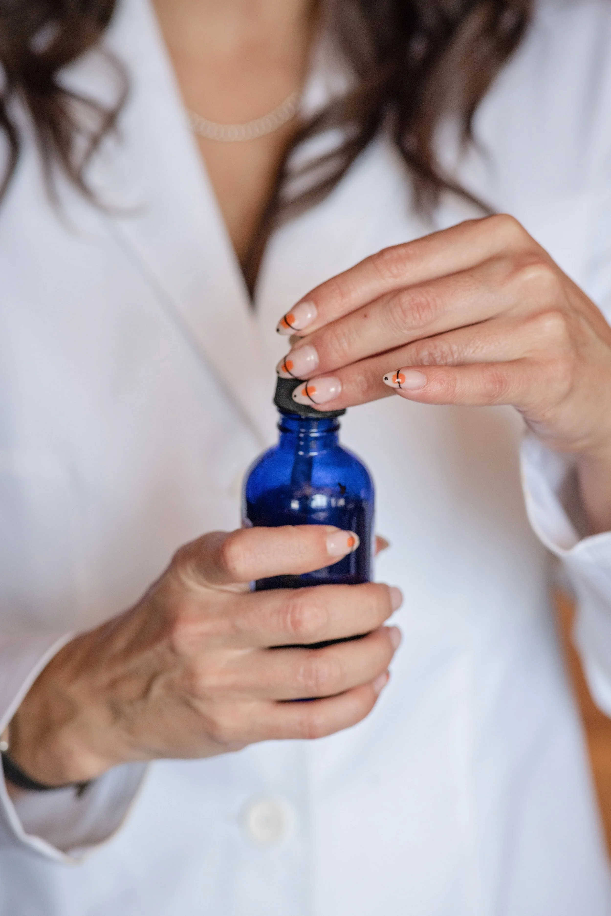 A woman with painted nails in orange and black, wearing a white coat and a pearl necklace, holding a small blue bottle with a dropper cap.