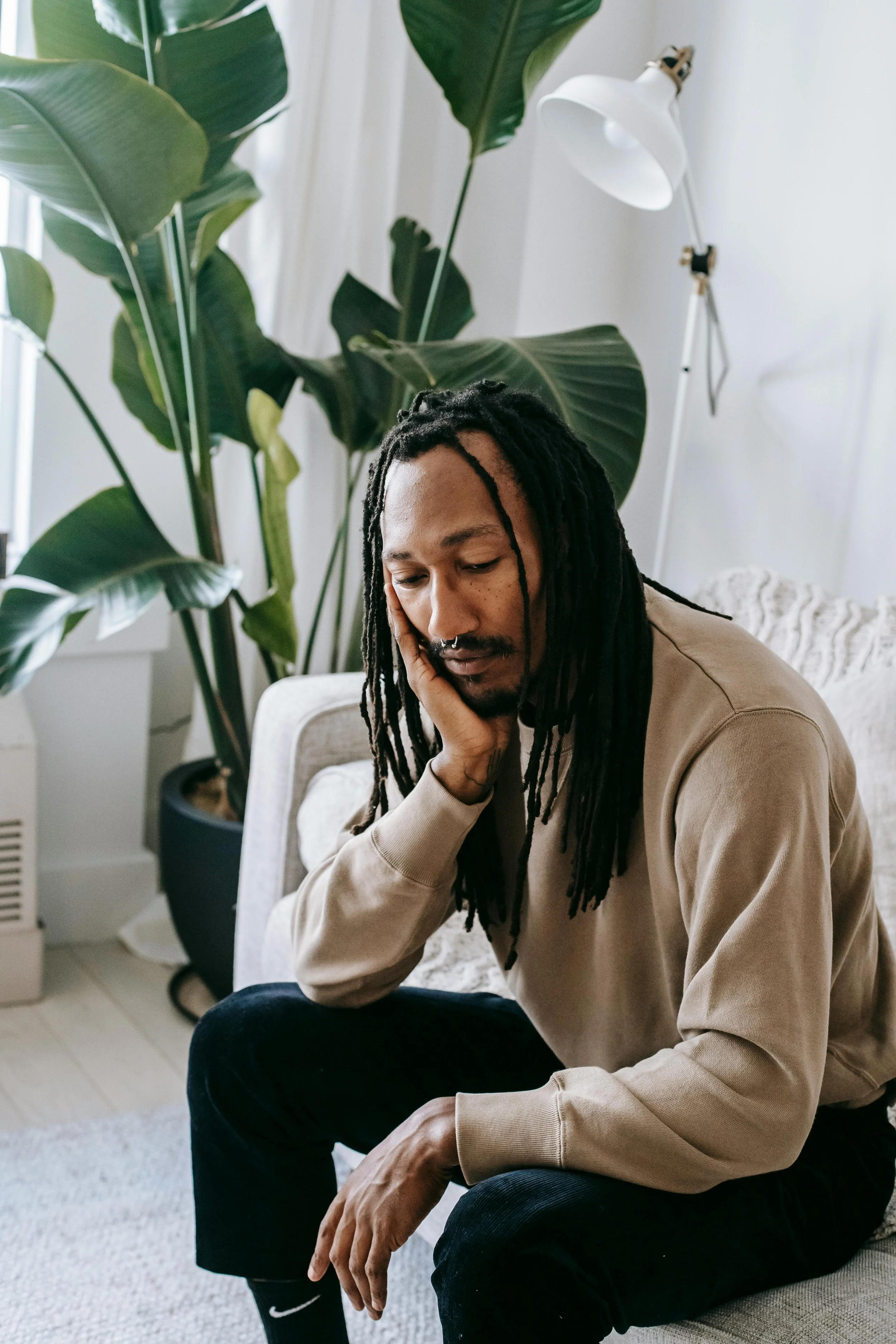 A man sitting on a sofa with his hand resting on his face, appearing contemplative or sad. The room has houseplants in the background and a floor lamp, with natural light coming through the window.
