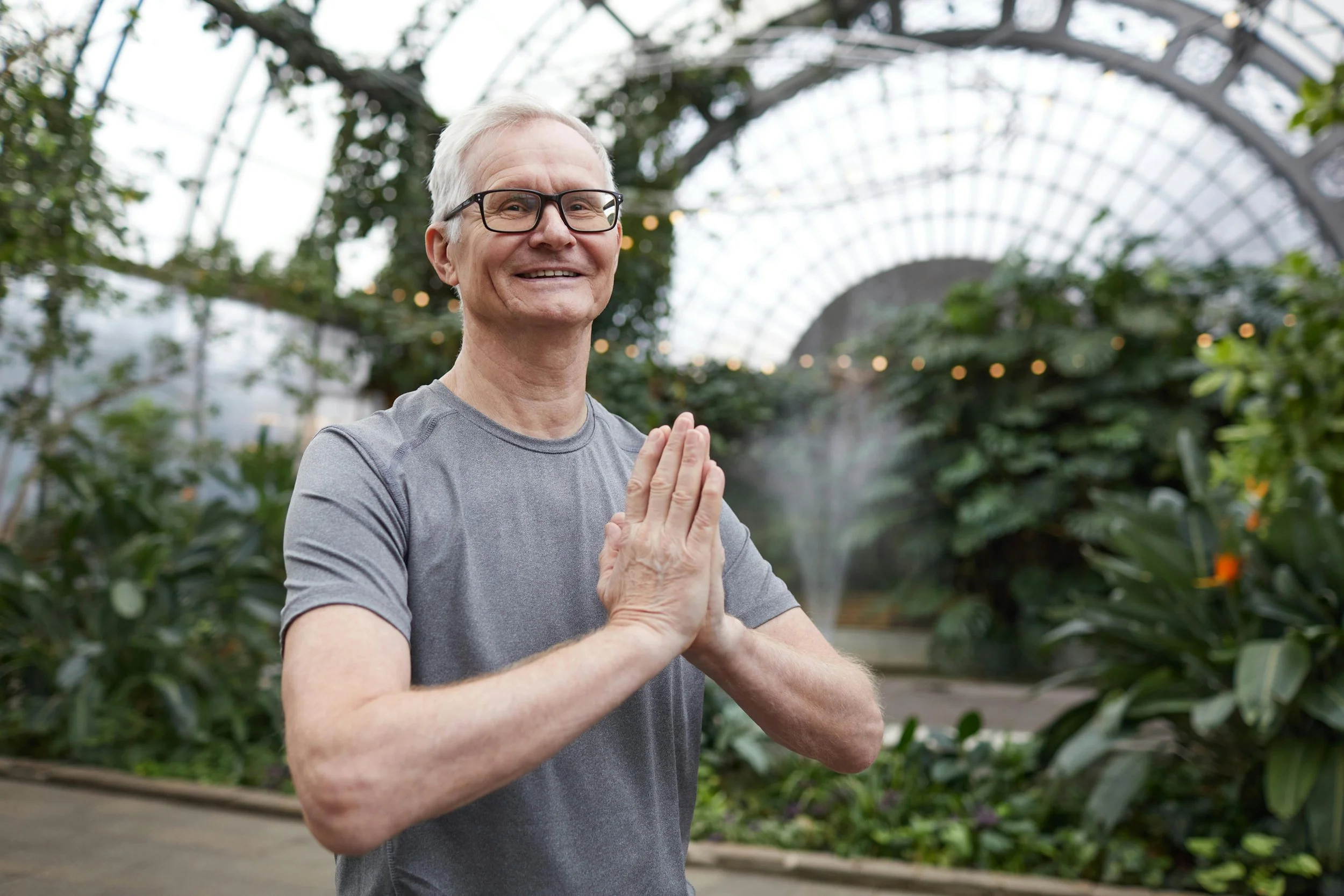 Older man smiling in a greenhouse, wearing glasses and a gray t-shirt, standing with his hands together in a prayer or meditation pose.
