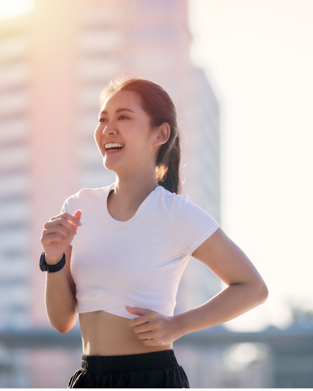 Young woman smiling and running outdoors in athletic clothes with city buildings in the background.