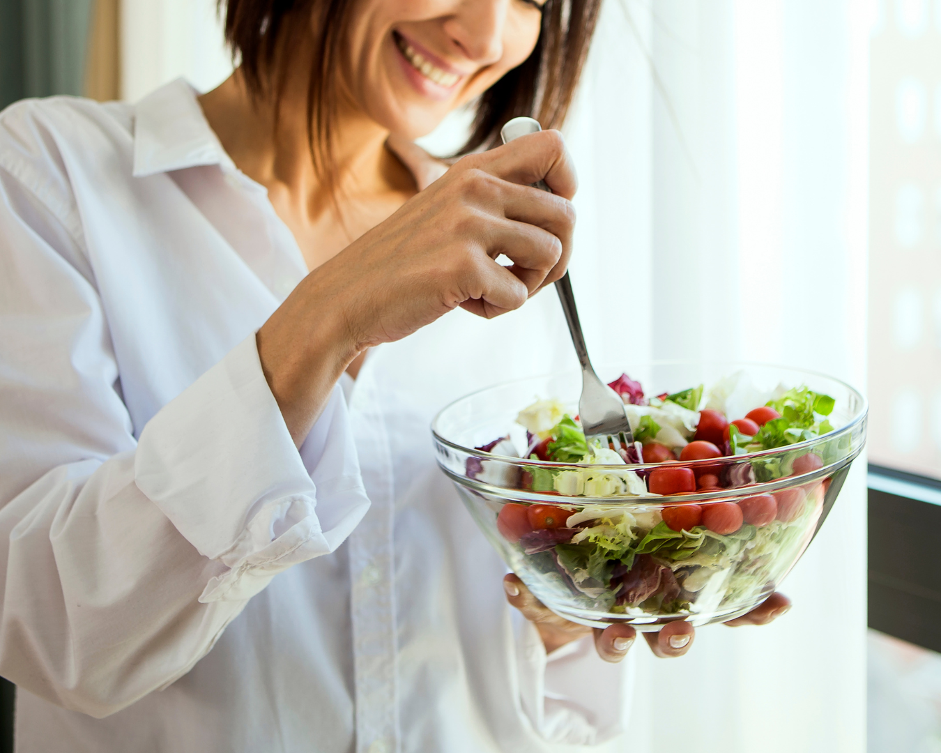 Person in white shirt smiling as they toss a fresh vegetable salad in a glass bowl.