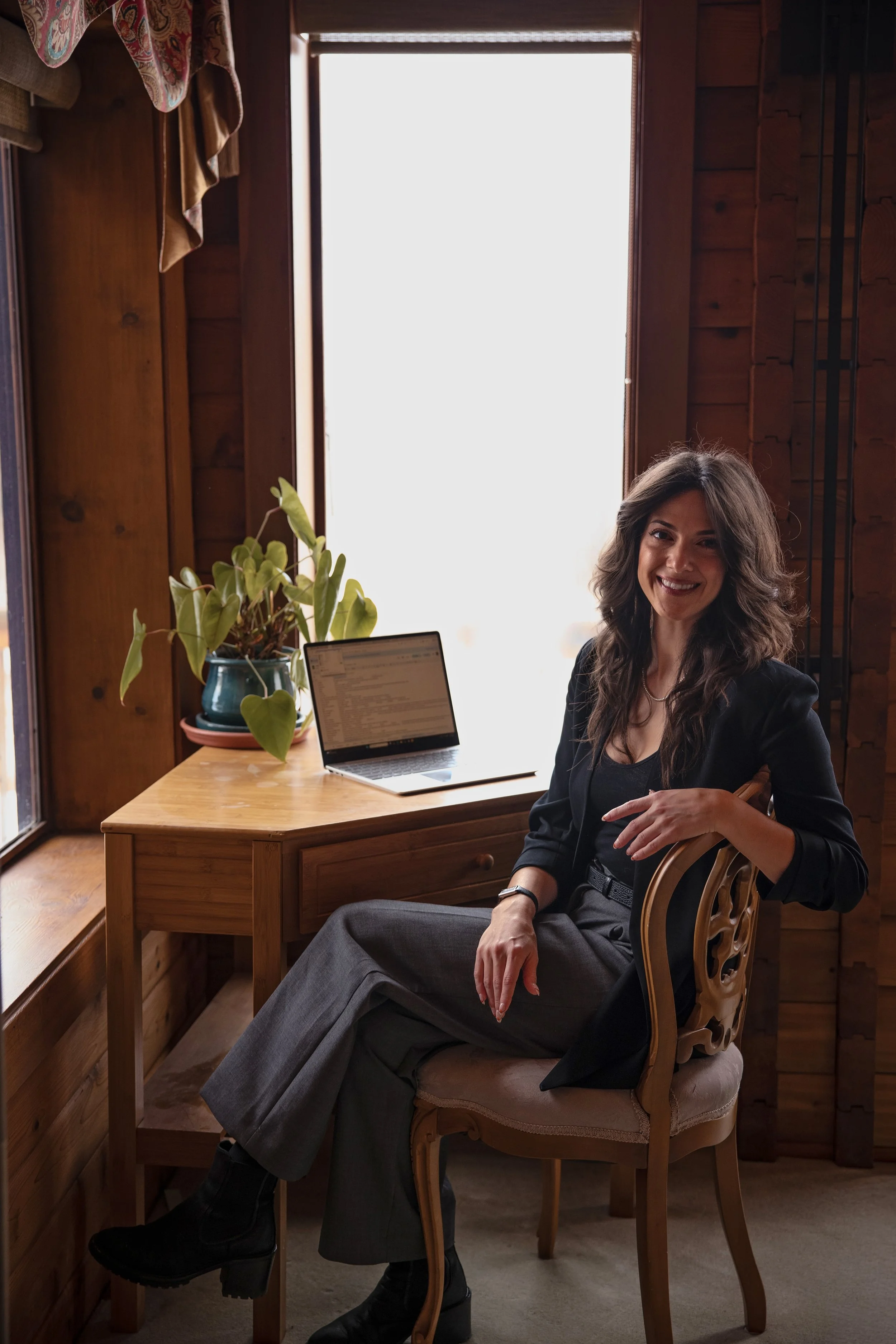 A woman with long wavy hair sitting on a wooden chair at a wooden desk near a window, smiling at the camera. The desk has a laptop and a potted plant.