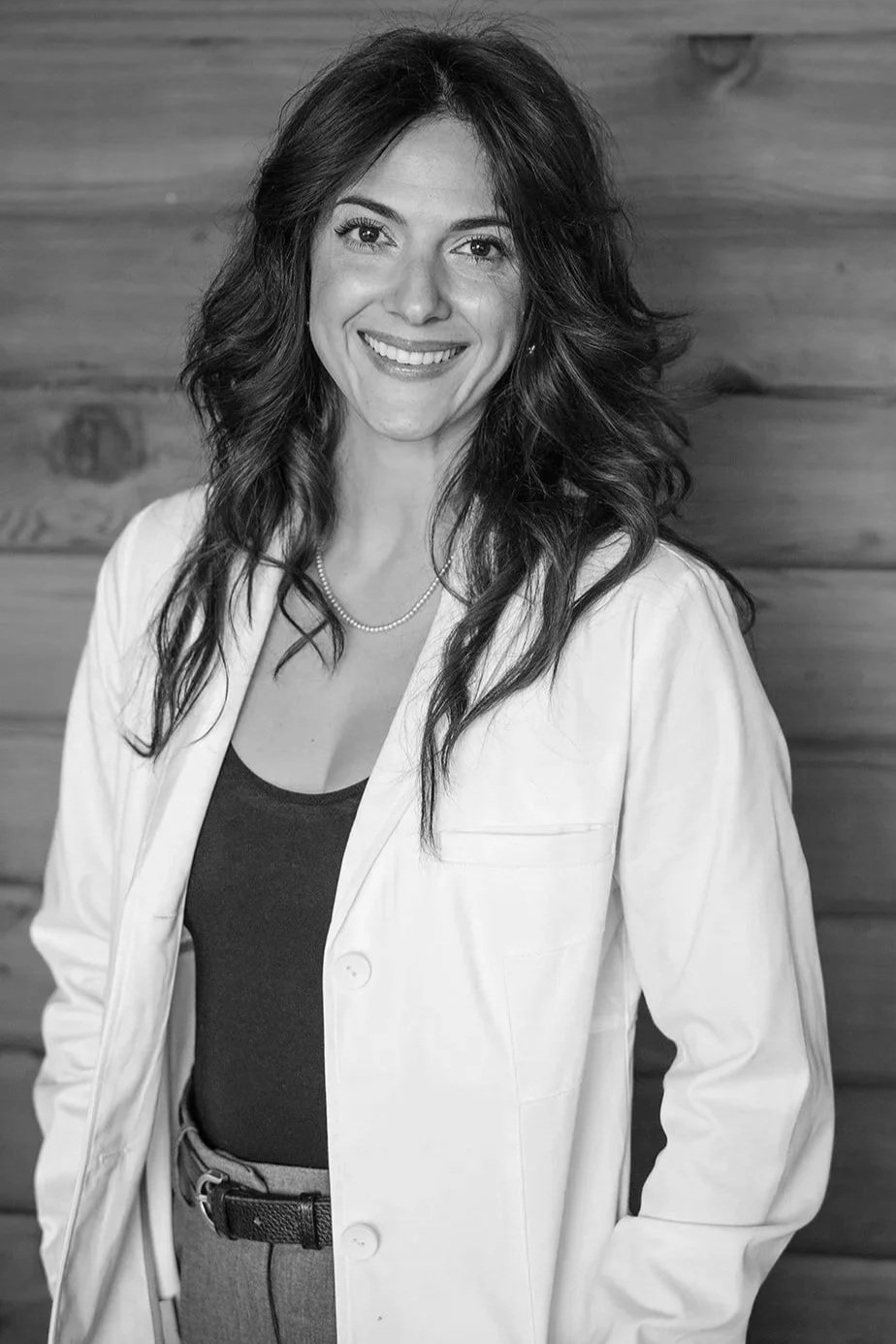 A woman with wavy dark hair, smiling, wearing a pearl necklace, a light-colored blazer, and dark top, standing in front of a wooden background.