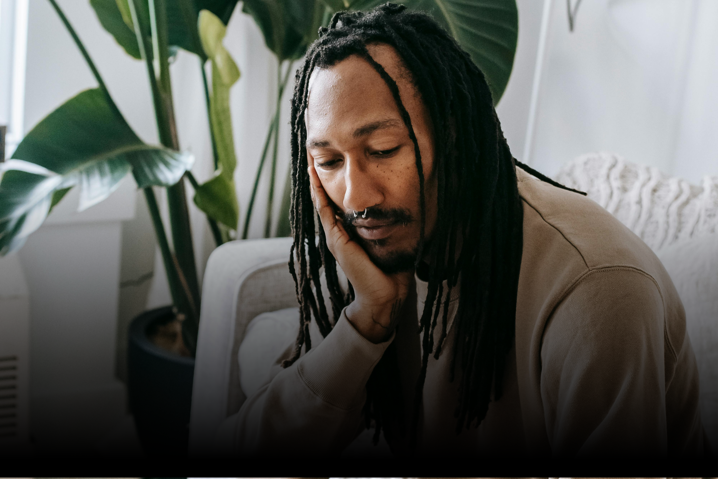 A man with long dreadlocks, a septum piercing, and a beard sitting indoors with his head resting on his hand, looking down with a somber expression, background includes large green indoor plants.
