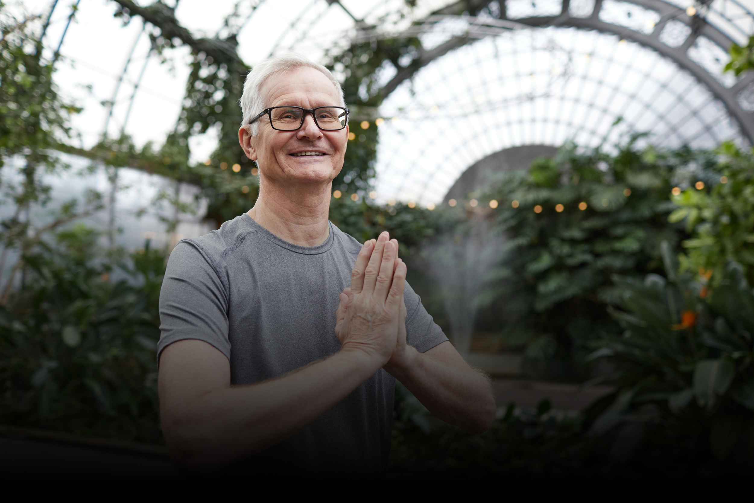 An elderly man with gray hair and glasses smiling and practicing yoga or meditation with hands in a prayer position in a greenhouse filled with plants and natural light.