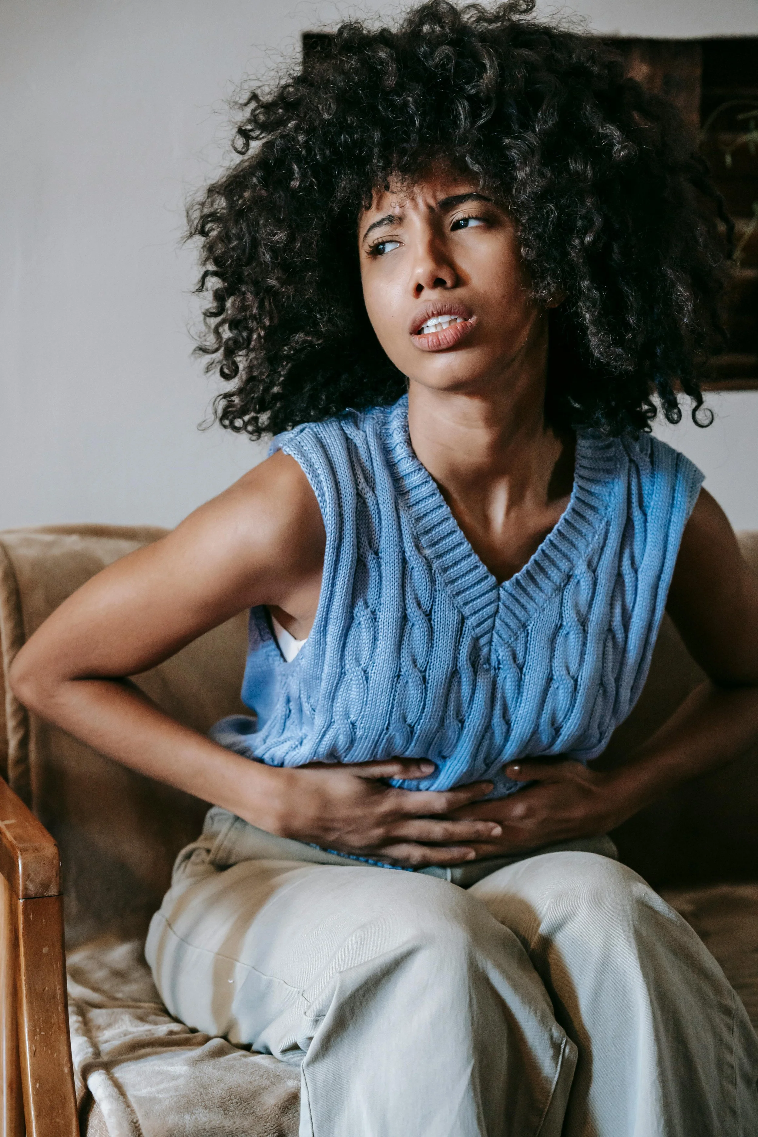 A woman with curly hair, wearing a blue sleeveless sweater, sitting on a beige couch, holding her stomach and appearing to experience stomach pain or discomfort.