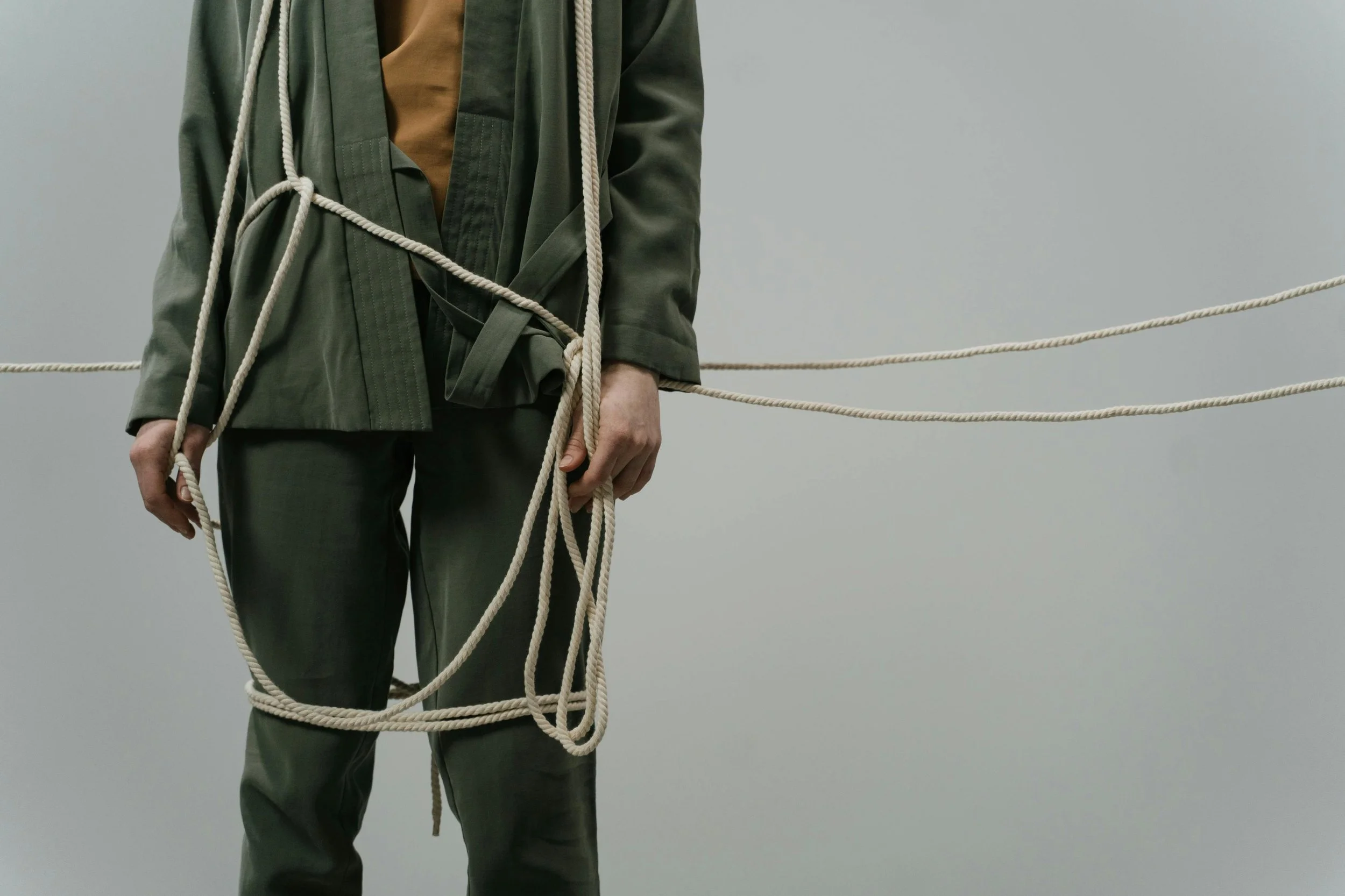 Person wearing dark green martial arts uniform with brown shirt underneath, surrounded by white ropes tied around their body and extending horizontally in both directions against a plain gray background.
