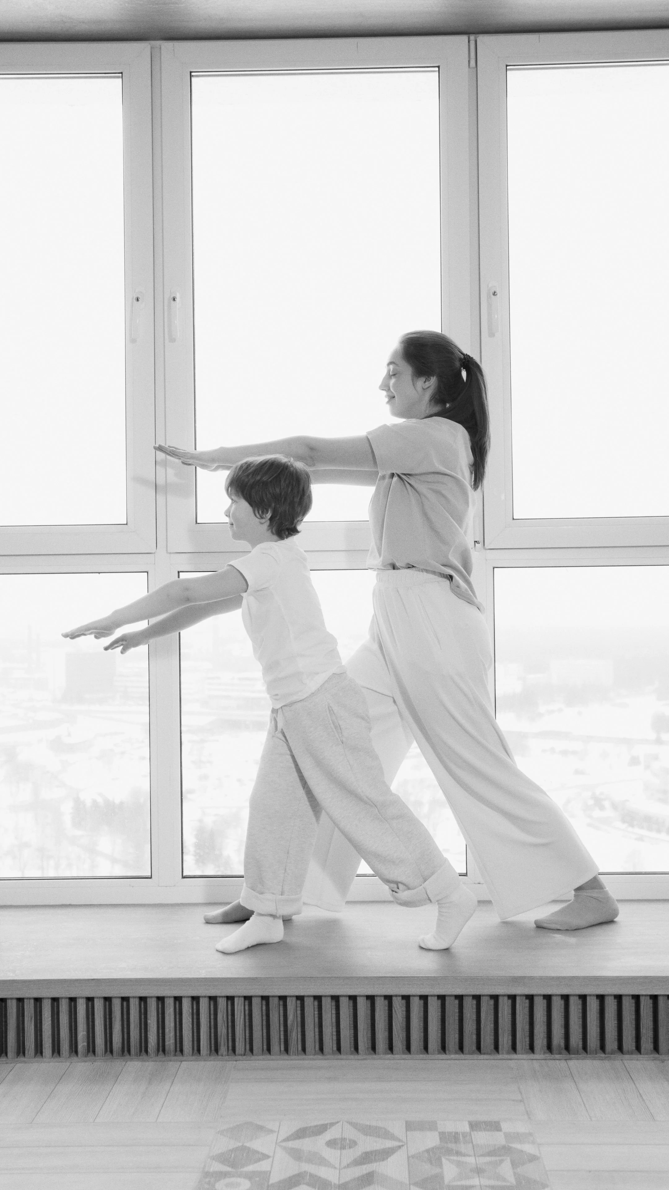 Une femme et un enfant faisant du yoga ensemble devant une grande fenêtre.