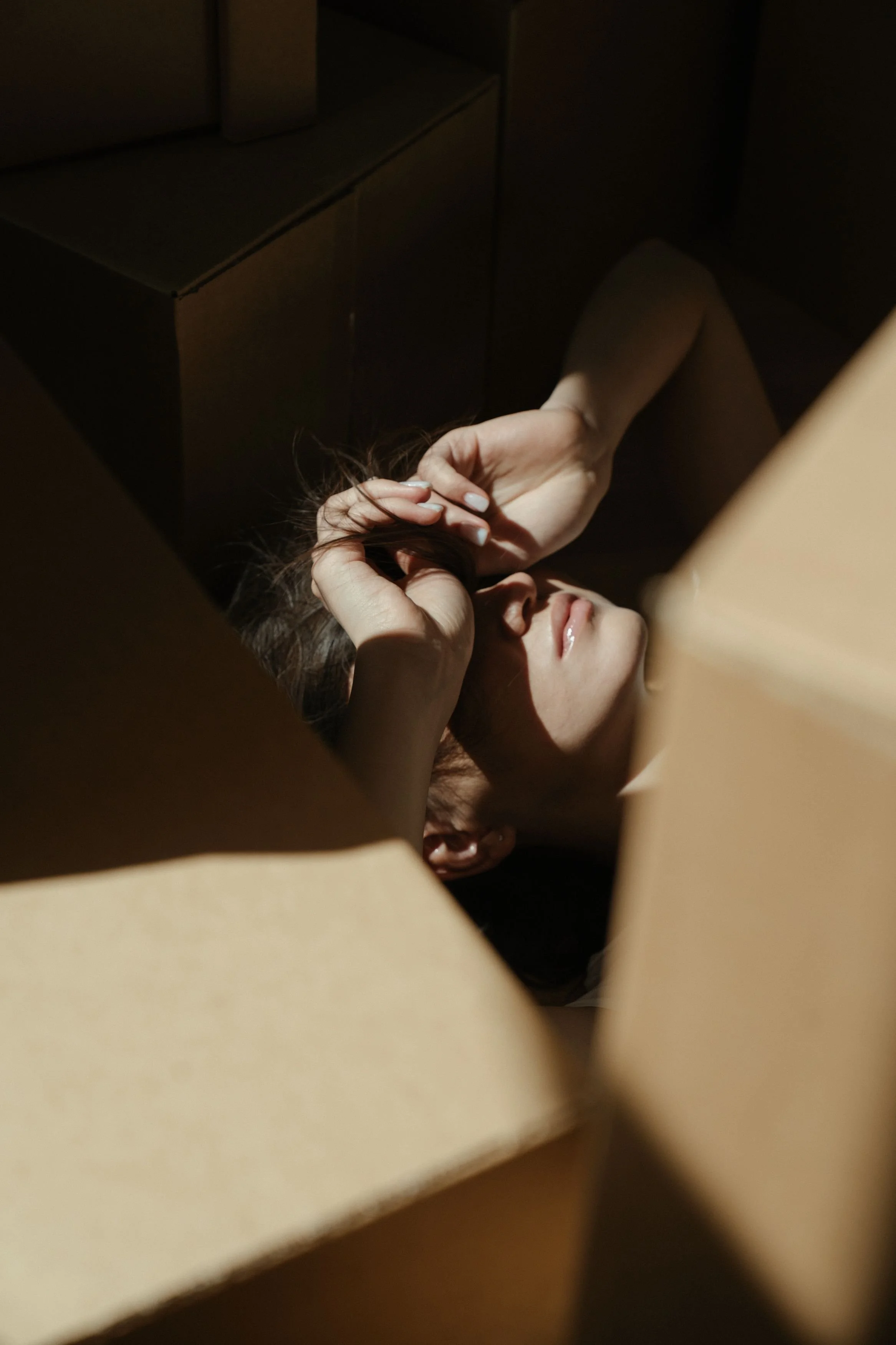 Une personne semble être allongée ou assise, entourée de murs ou de meubles en carton ou en bois, créant une ambiance tamisée ou sombre.