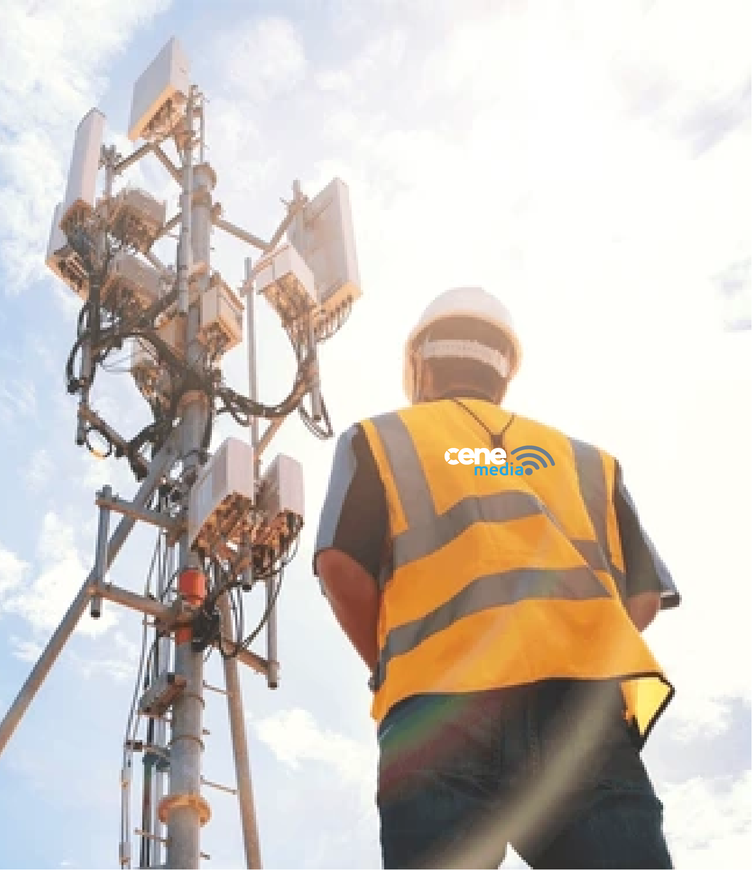 Worker in a yellow safety vest and helmet inspecting a cell tower under a bright, partly cloudy sky.