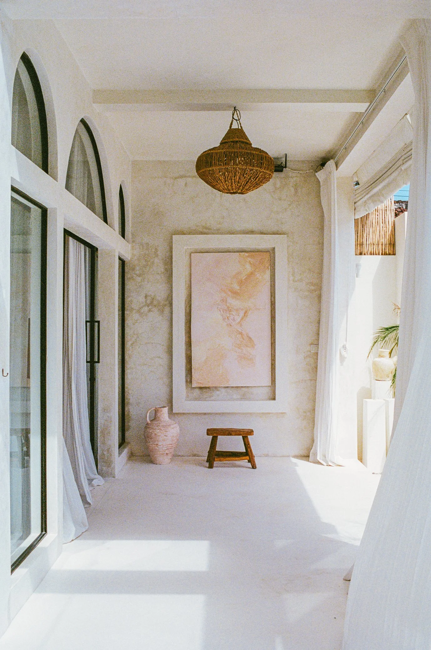 Bright, minimalist room with white walls and floor, a large framed abstract painting, a small wooden stool, a pottery vase, and white curtains. Natural light streams through arched windows and an open door.
