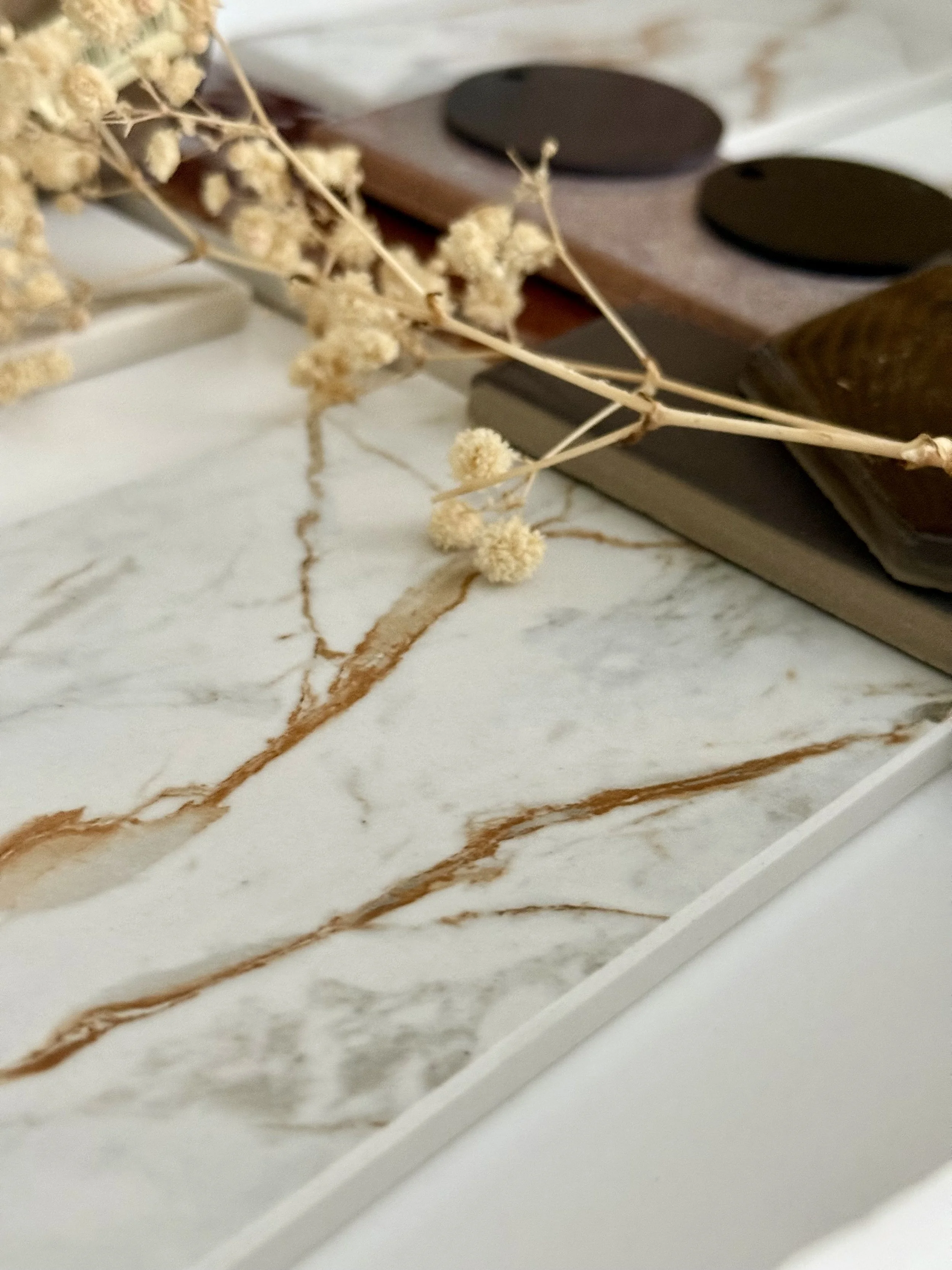 Close-up of a white and brown marble surface with dried beige flowers and dark-colored round objects arranged on top.