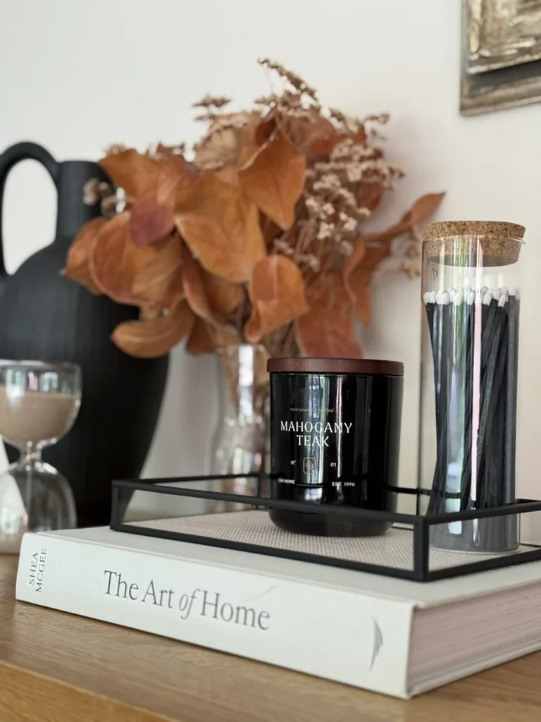 Styled console vignette featuring a black vase, autumn foliage, candle, decorative matches and coffee table books in warm earthy tones.