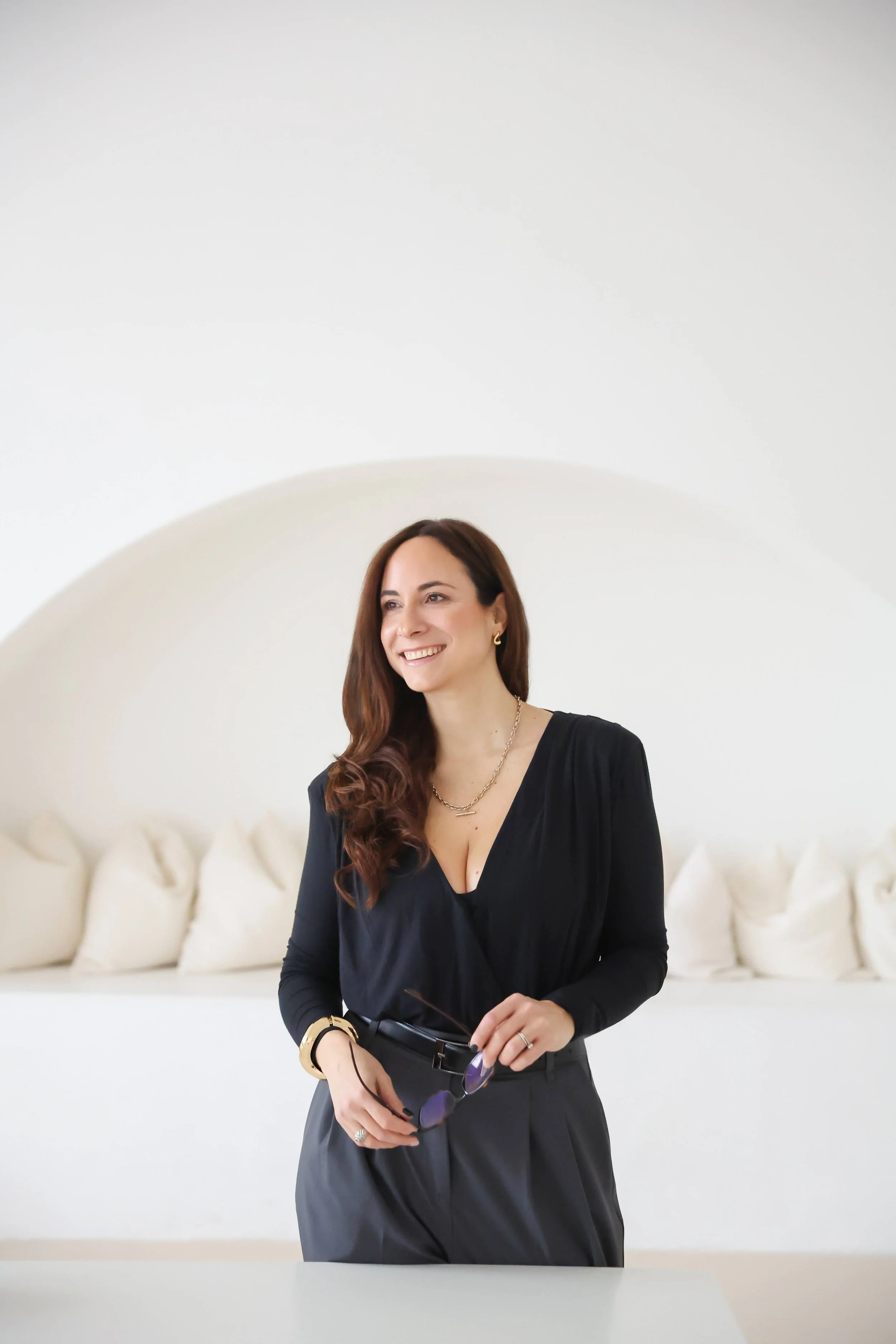 Fed stands in the centre of the image wearing all black and gold jewellery. She is smiling and looking off to the left of the camera. Behind her, is a beautiful studio with white arched walls and cream cushions. The photo feels light and bright.