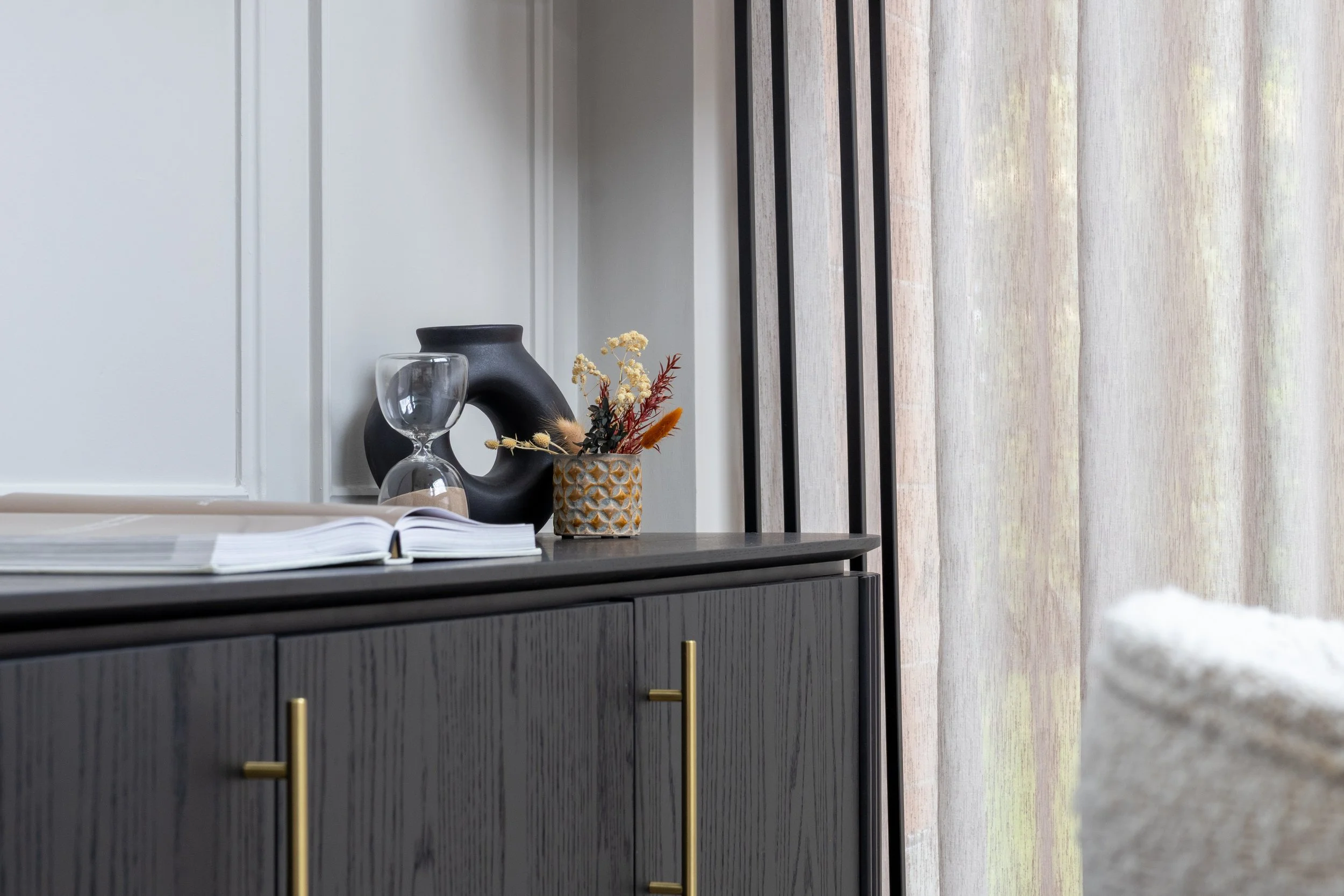 Grey cupboard units with gold handles are the main focal point of the picture. On top, sites a black circular vase, a coffee-table book that's open to the middle, and a patterned ceramic plant pot filled with small leaves