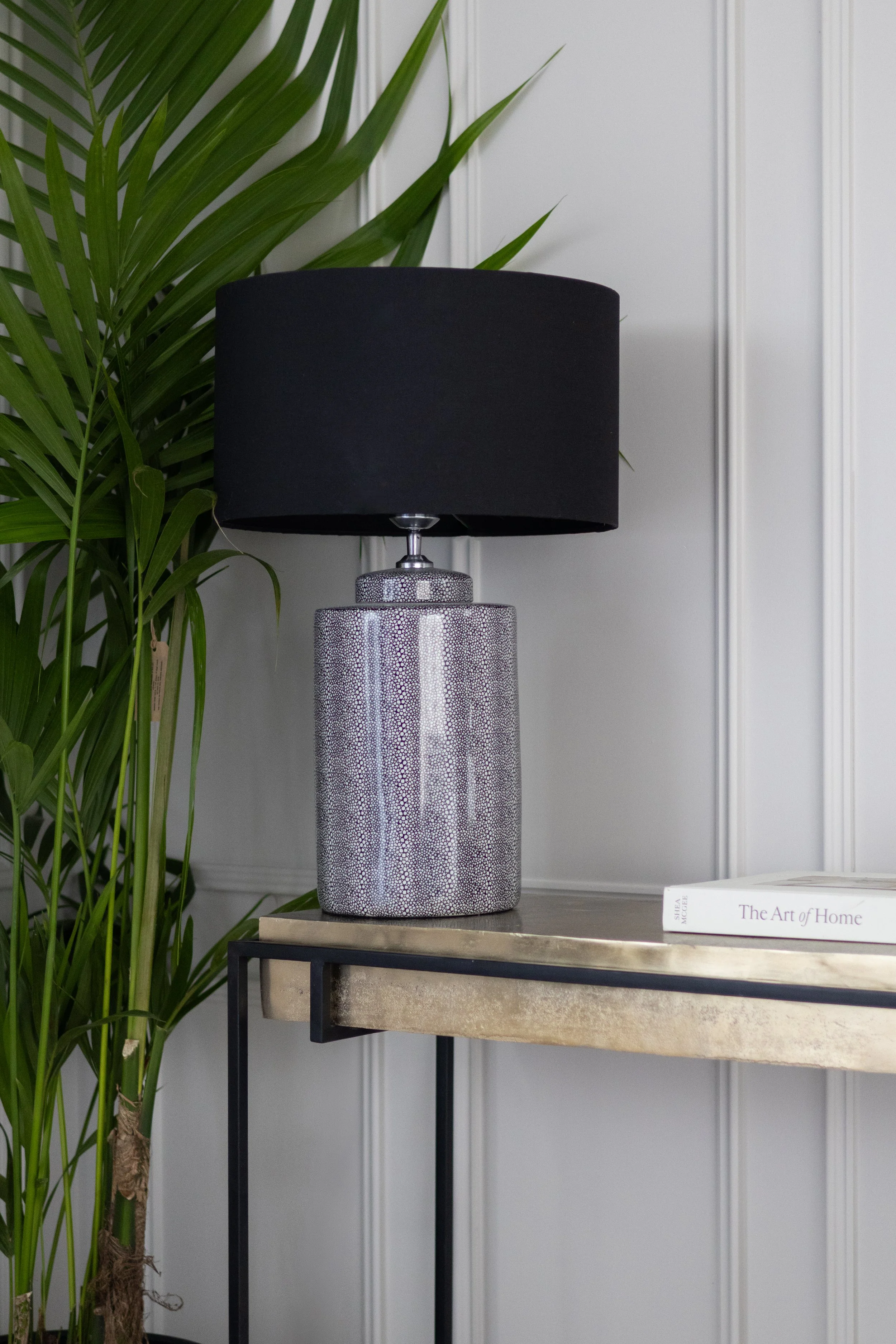 A decorative room corner featuring a textured silver table lamp with a black lampshade, placed on a small table with a metal frame and marble surface, with a large green potted plant and a white book titled 'The Art of Home' beside it.