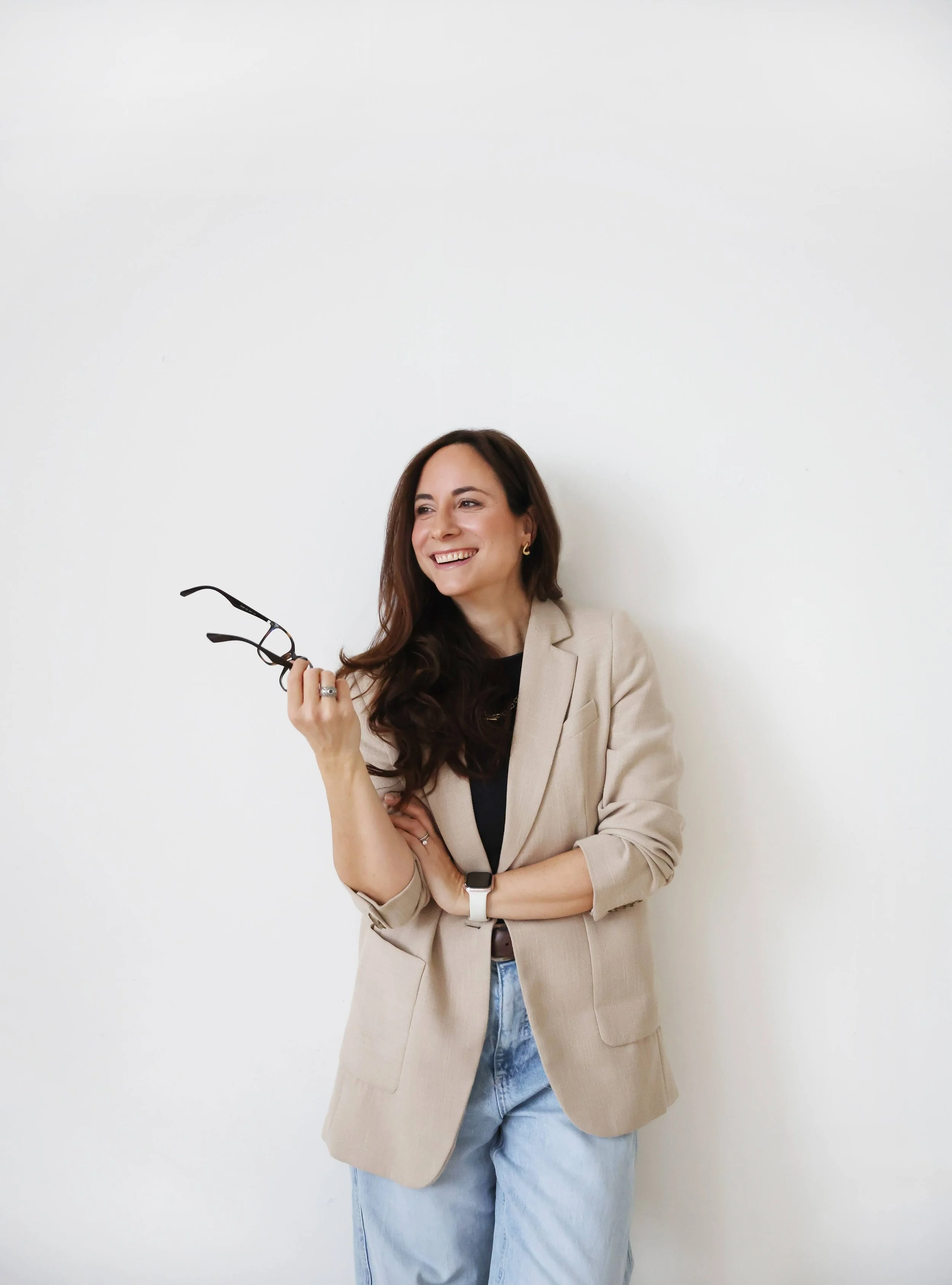 Fed stands against a white wall in a neutral, oversized blazer, a black top and light-blue jeans. She's looking off to the left, smiling with her glasses in her hand. The photo feels bright and joyful.