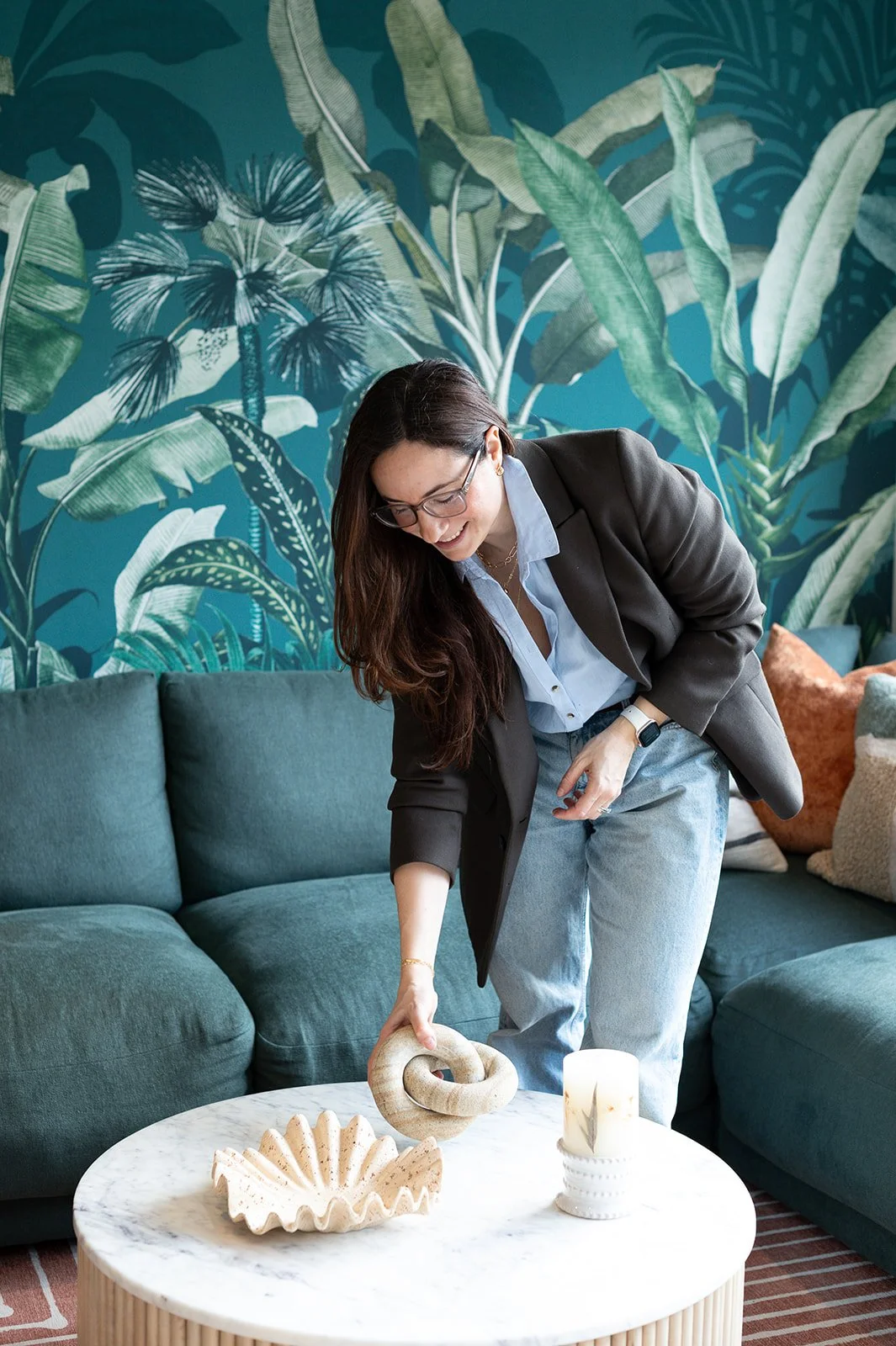 Fed, wearing a dark suit jacket, a blue striped shirt and blue jeans is decorating a coffee table with a ceramic knot sculpture, a shell-shaped dish, and a lit candle, in a living room with botanical murals on the wall.