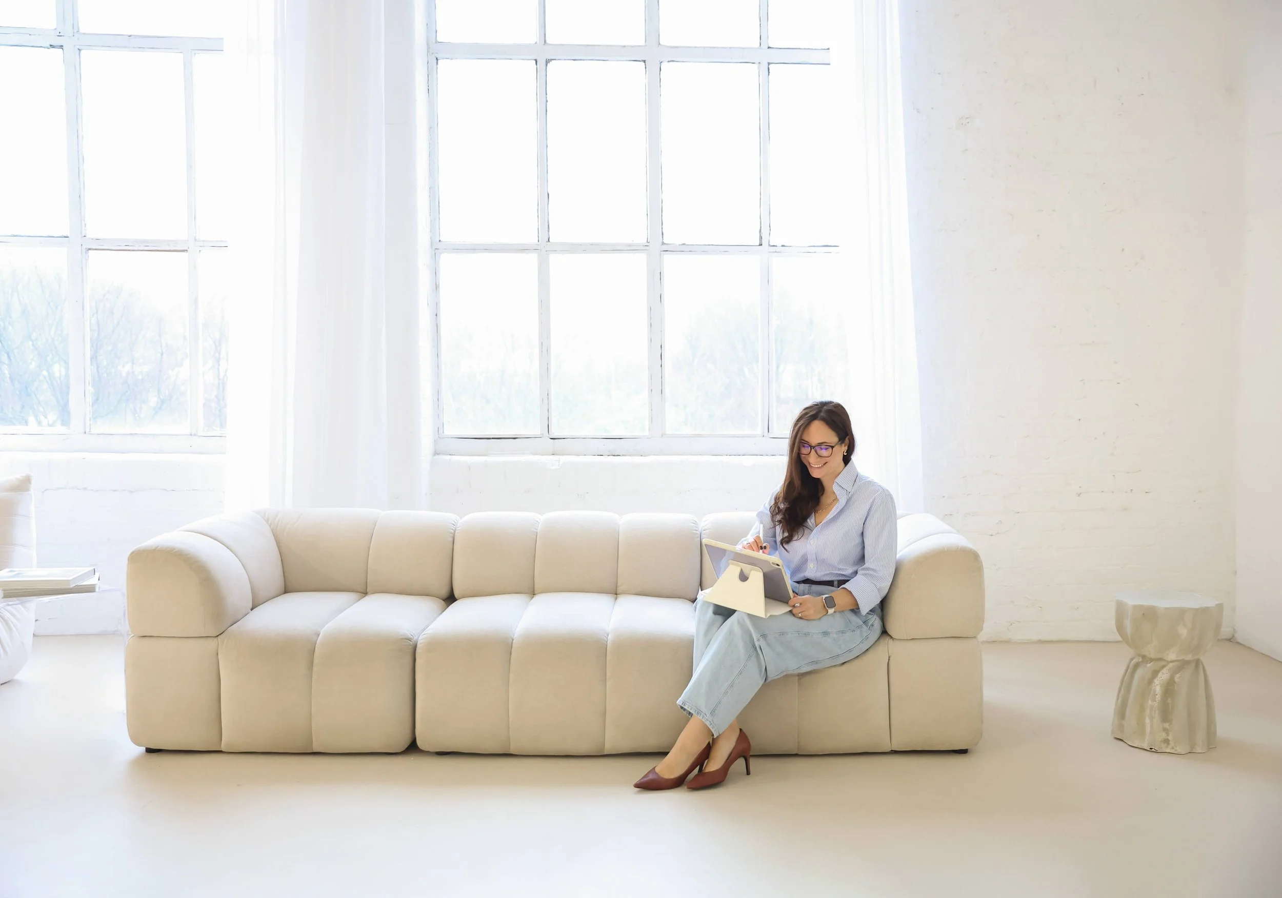Fed sits on a cream-coloured sofa, using a tablet in a bright, modern room with large windows and white walls, wearing glasses, a light blue button-up shirt, light-coloured pants, and high heels.