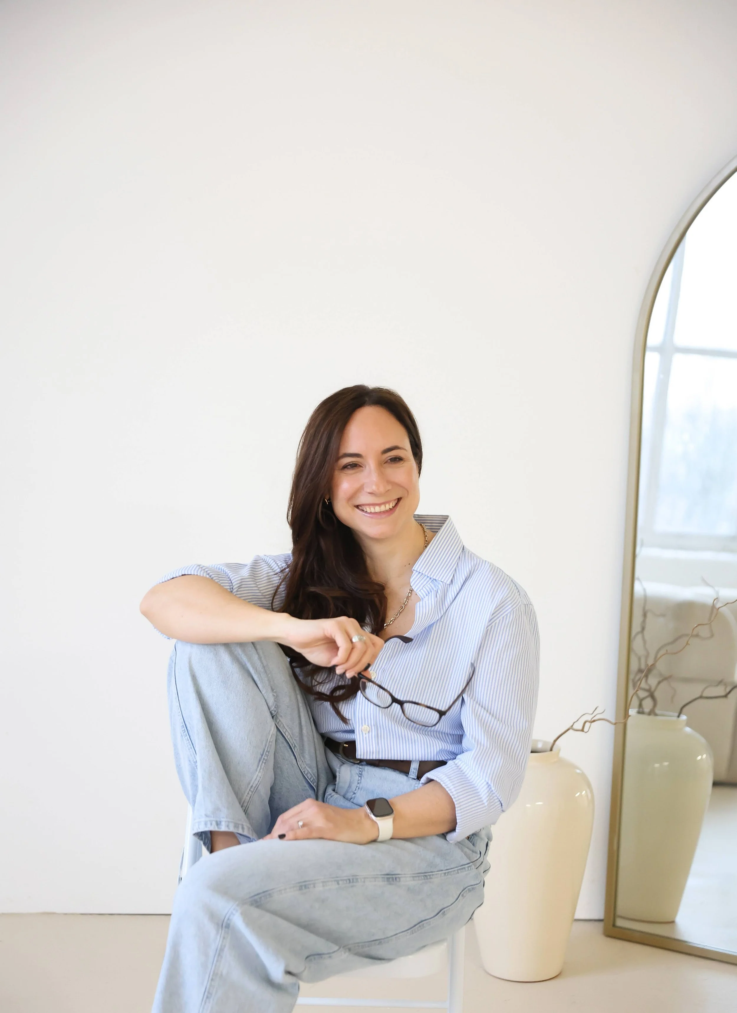 Fed sits on a chair in a casual position wearing a blue striped shirt and light-blue jeans. She is smiling and looking just off to the right of the camera holding her glasses in her hand. Behind her is a organic plant pot and floor-length mirror.