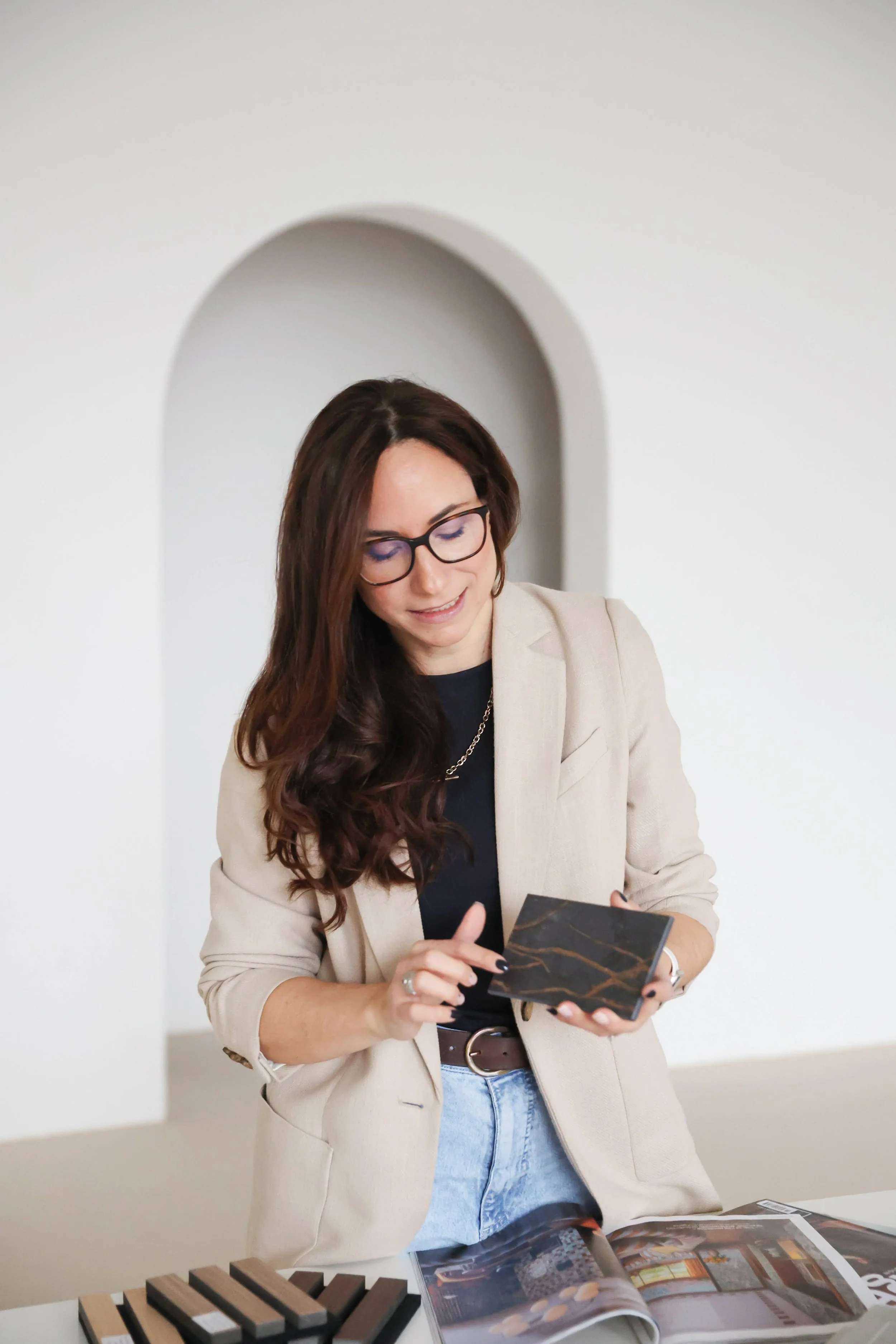 Fed, wearing her glasses, a beige blazer, and a black shirt, stands at a table with photo samples and holding a booklet or brochure with a marble pattern cover.