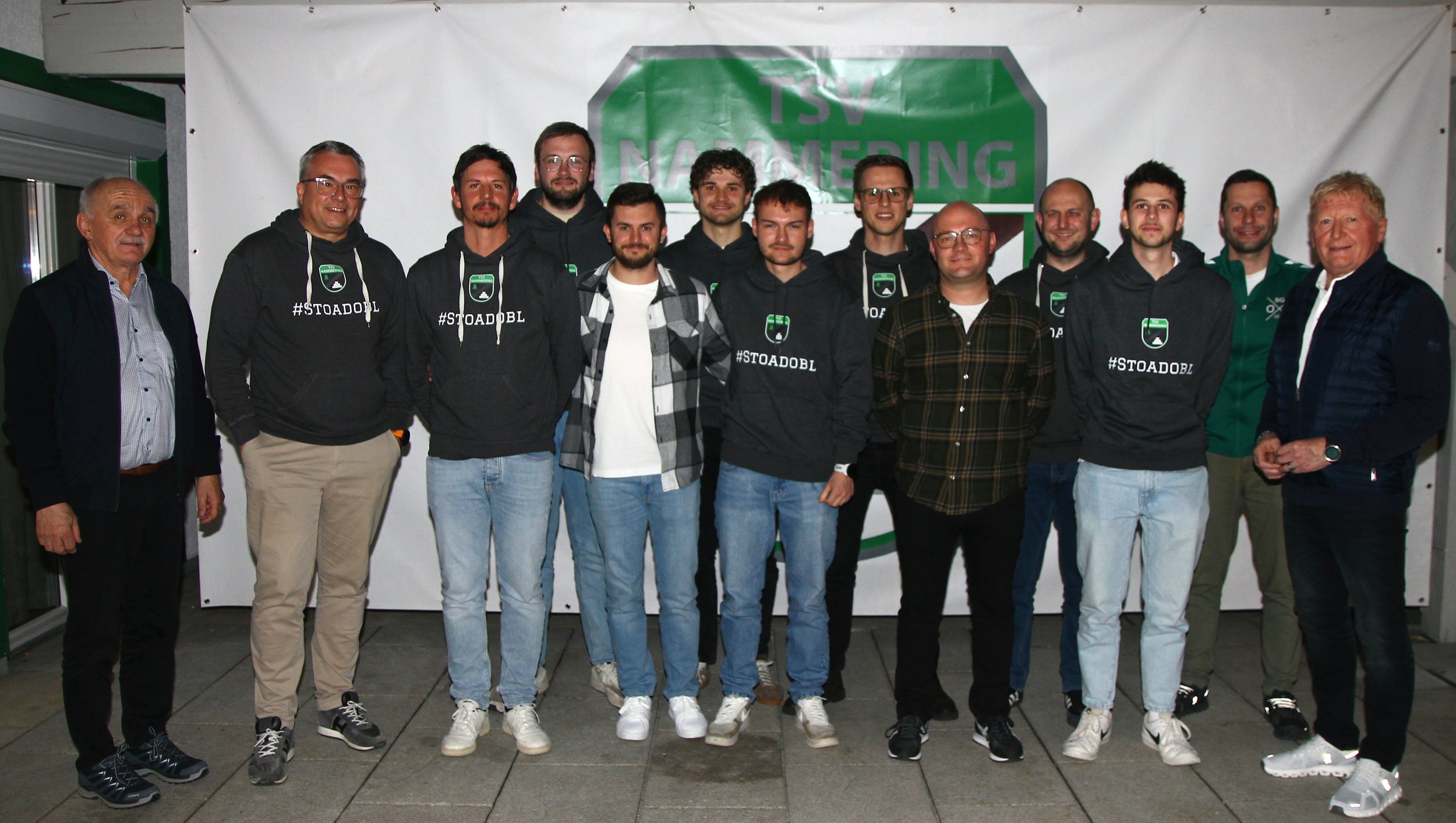 Gruppenfoto von mehreren Männern, einige tragen schwarze Hoodies mit einem Logo und Hashtag #STOADOBL, vor einem Banner mit dem Schriftzug TSV, vermutlich in einer Sporthalle oder einem Veranstaltungsraum.