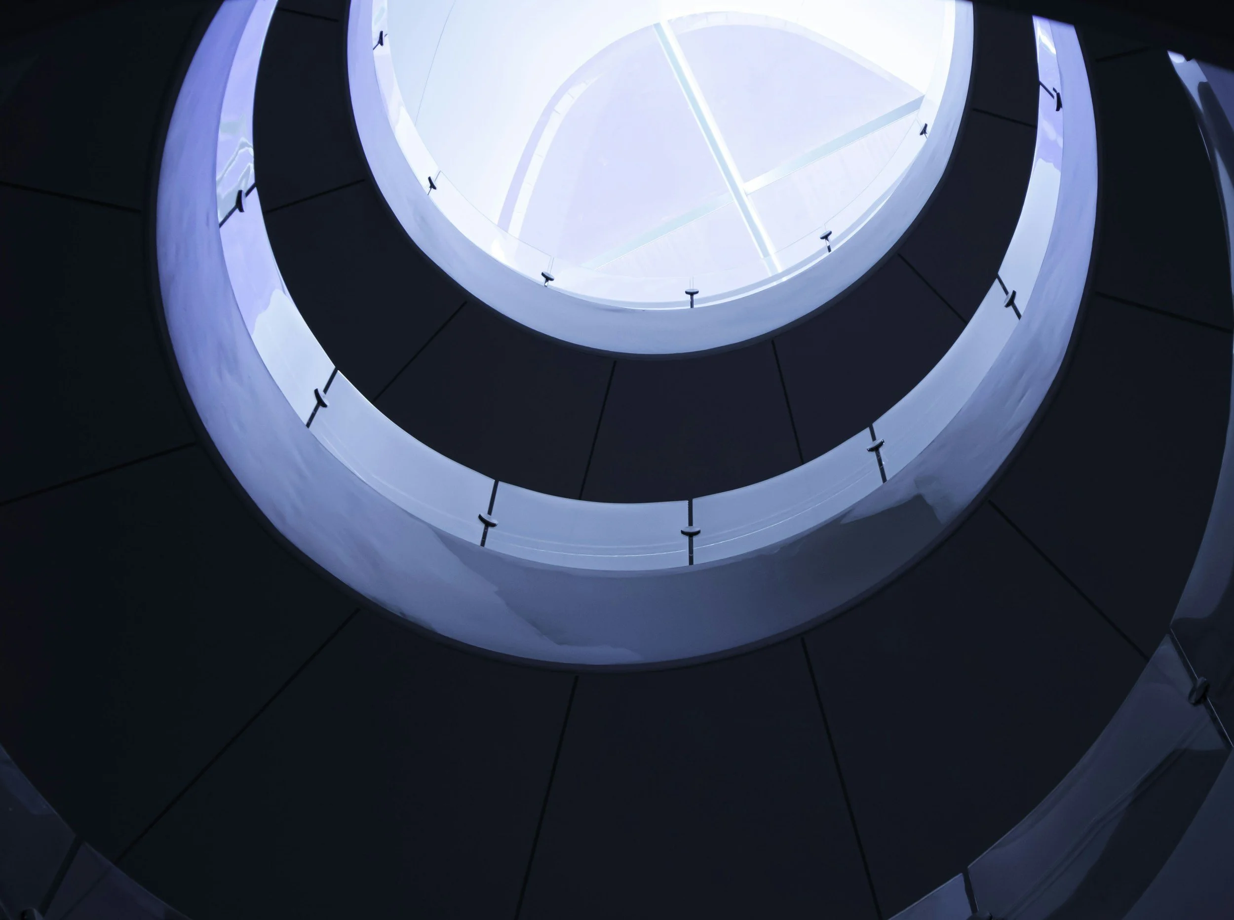 Looking up at a modern glass spiral staircase with a circular opening at the top, metal railings, and sleek dark walls.