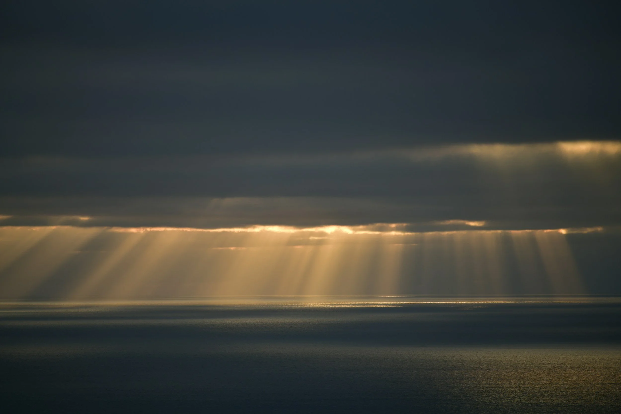 Sun rays breaking through dark clouds over a body of water during sunset or sunrise.