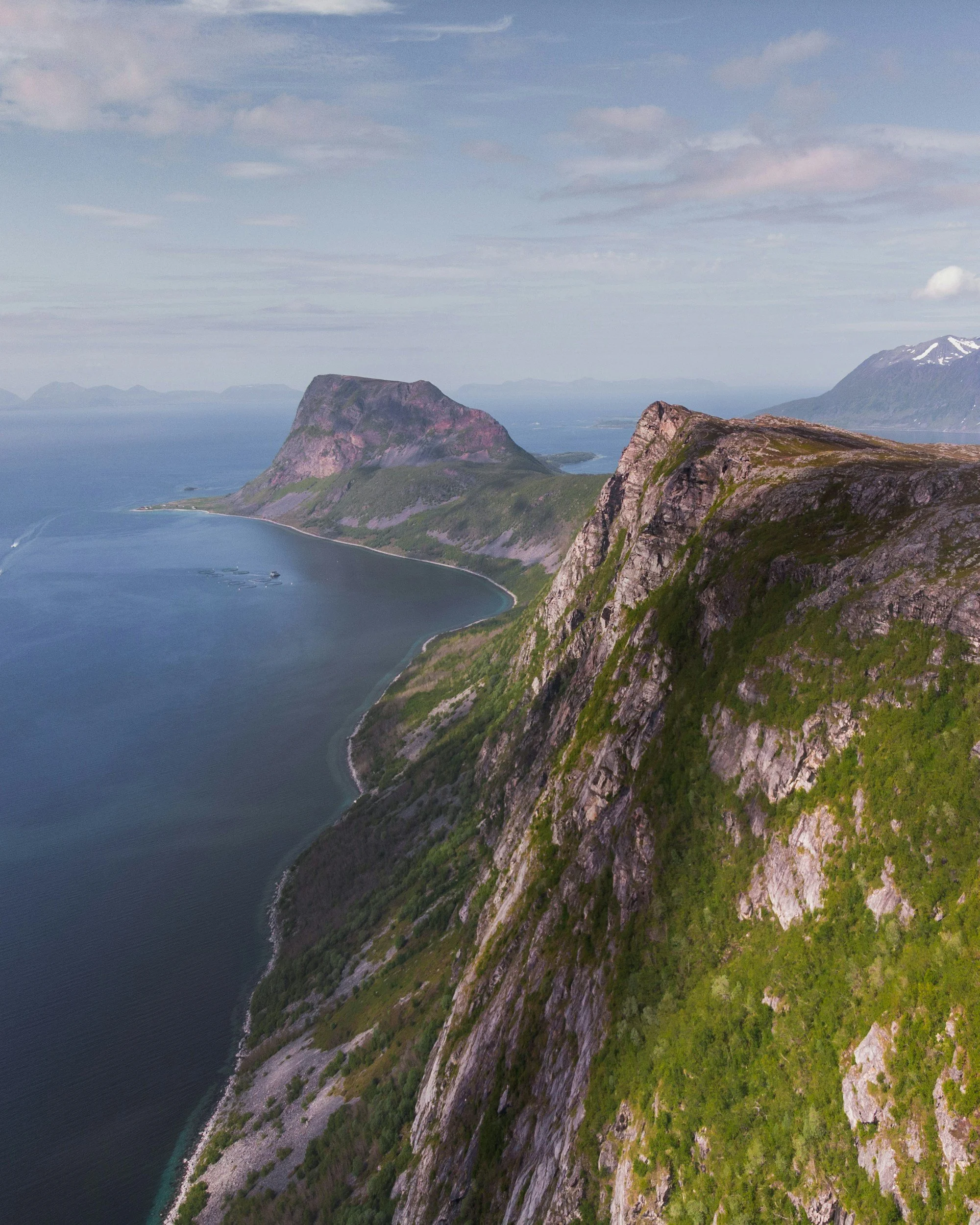 Aerial view of rugged island cliffs with green vegetation, overlooking a calm body of water with a small boat, mountains in the background, and partly cloudy sky.