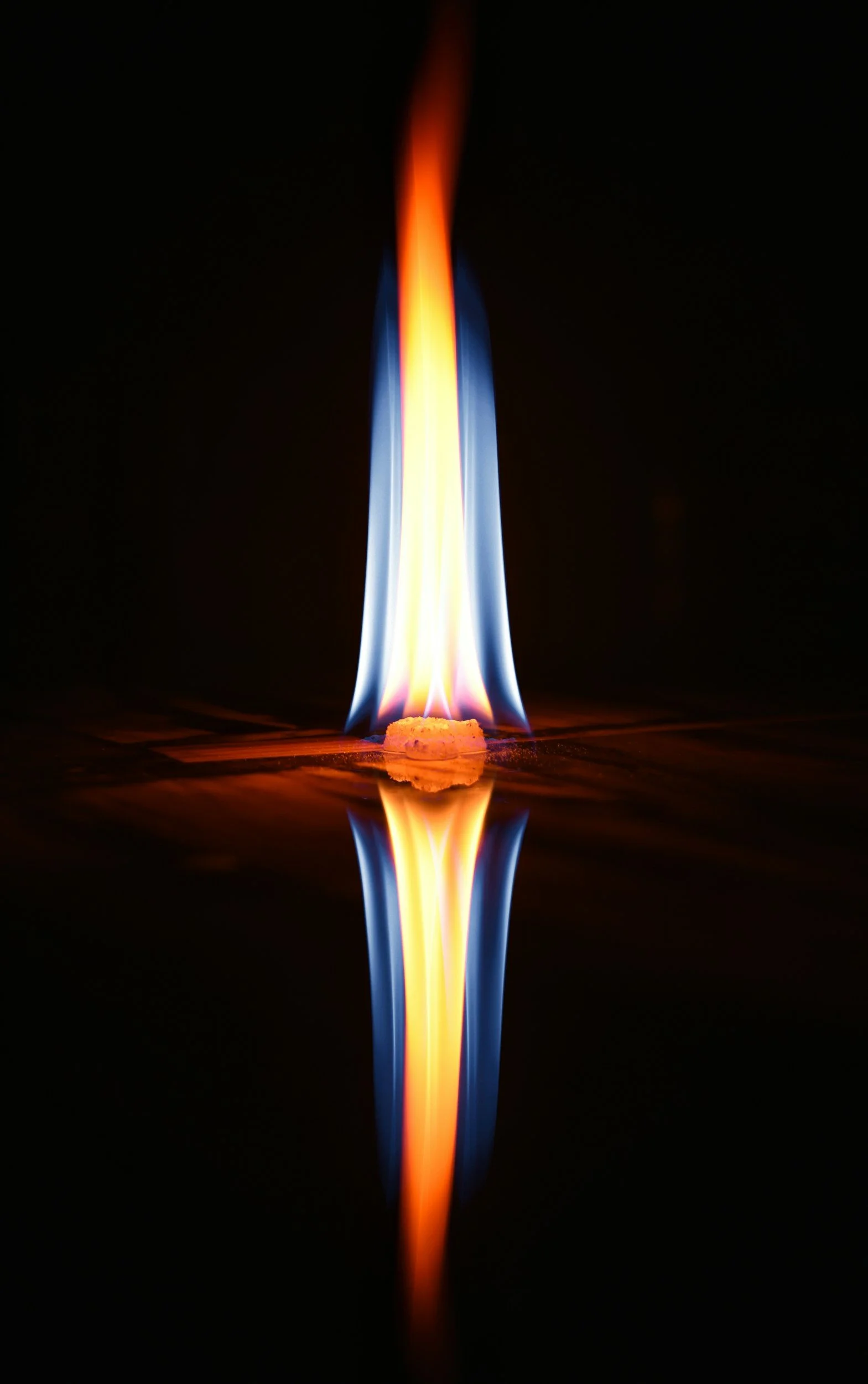 A close-up of a blue and orange flame burning with its reflection visible on a dark surface.