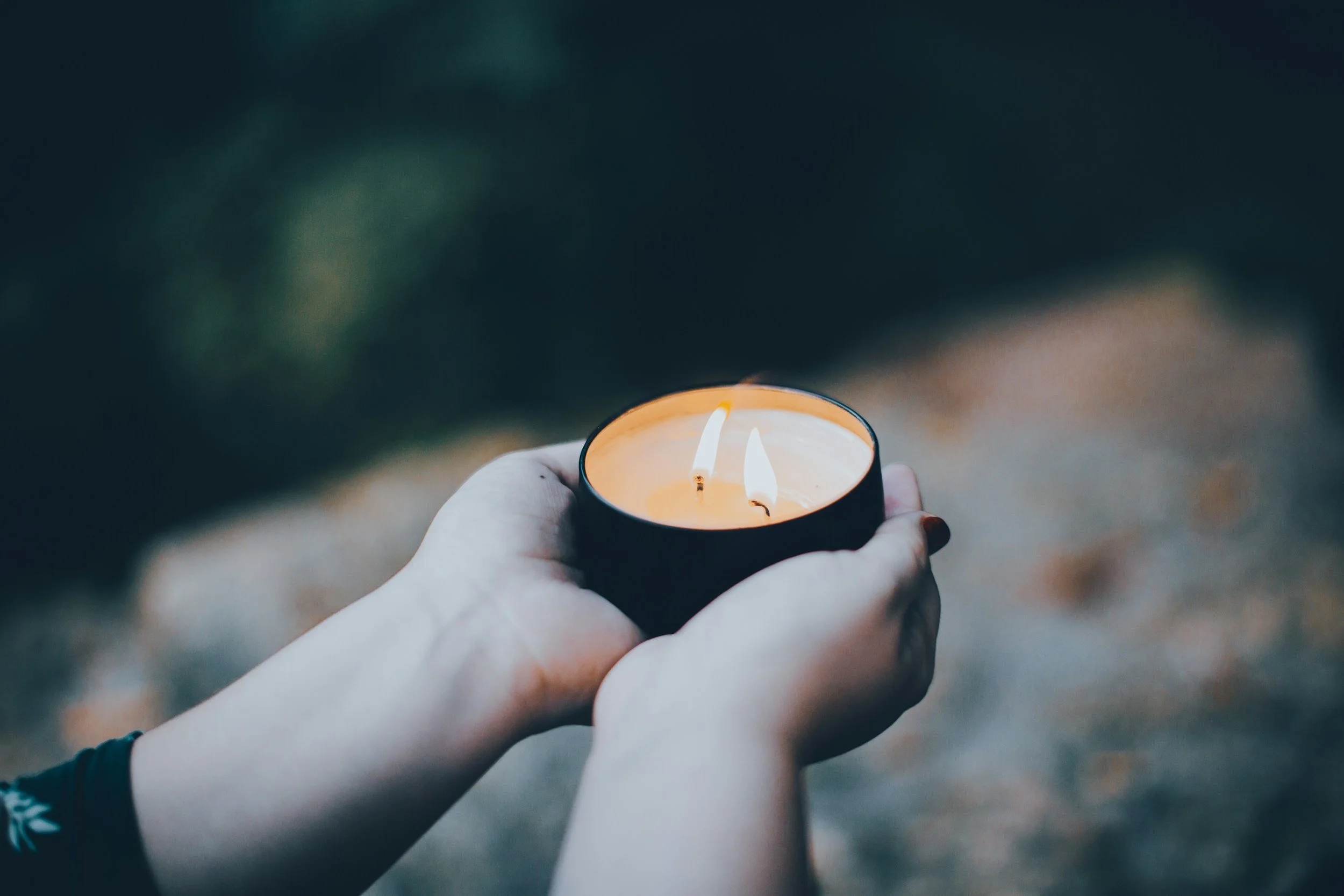 Hands holding a lit candle in a black holder outdoors at dusk.