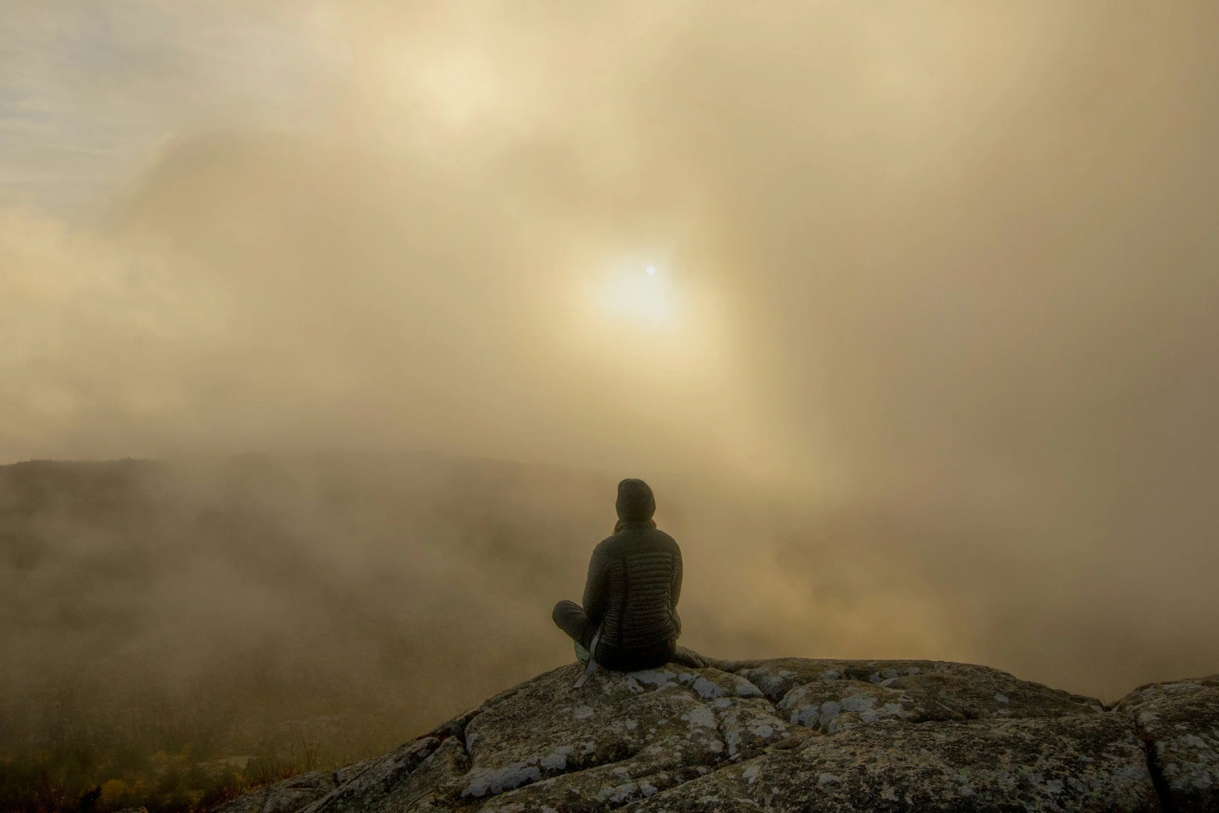 A person sitting on a rock overlooking a misty landscape during sunrise or sunset.