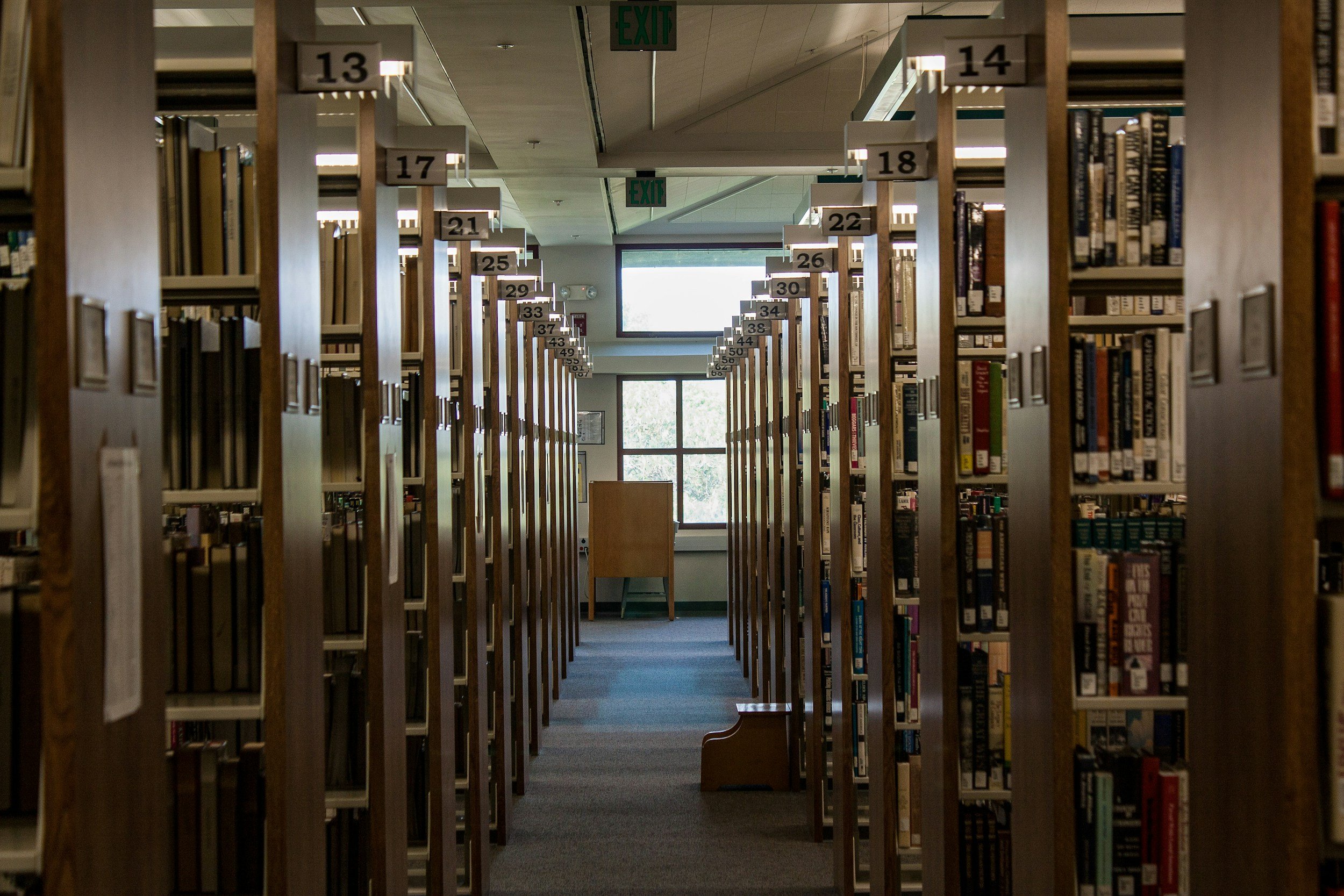 View of an aisle in a library with rows of bookshelves labeled with numbers. A window at the end of the aisle lets in natural light.