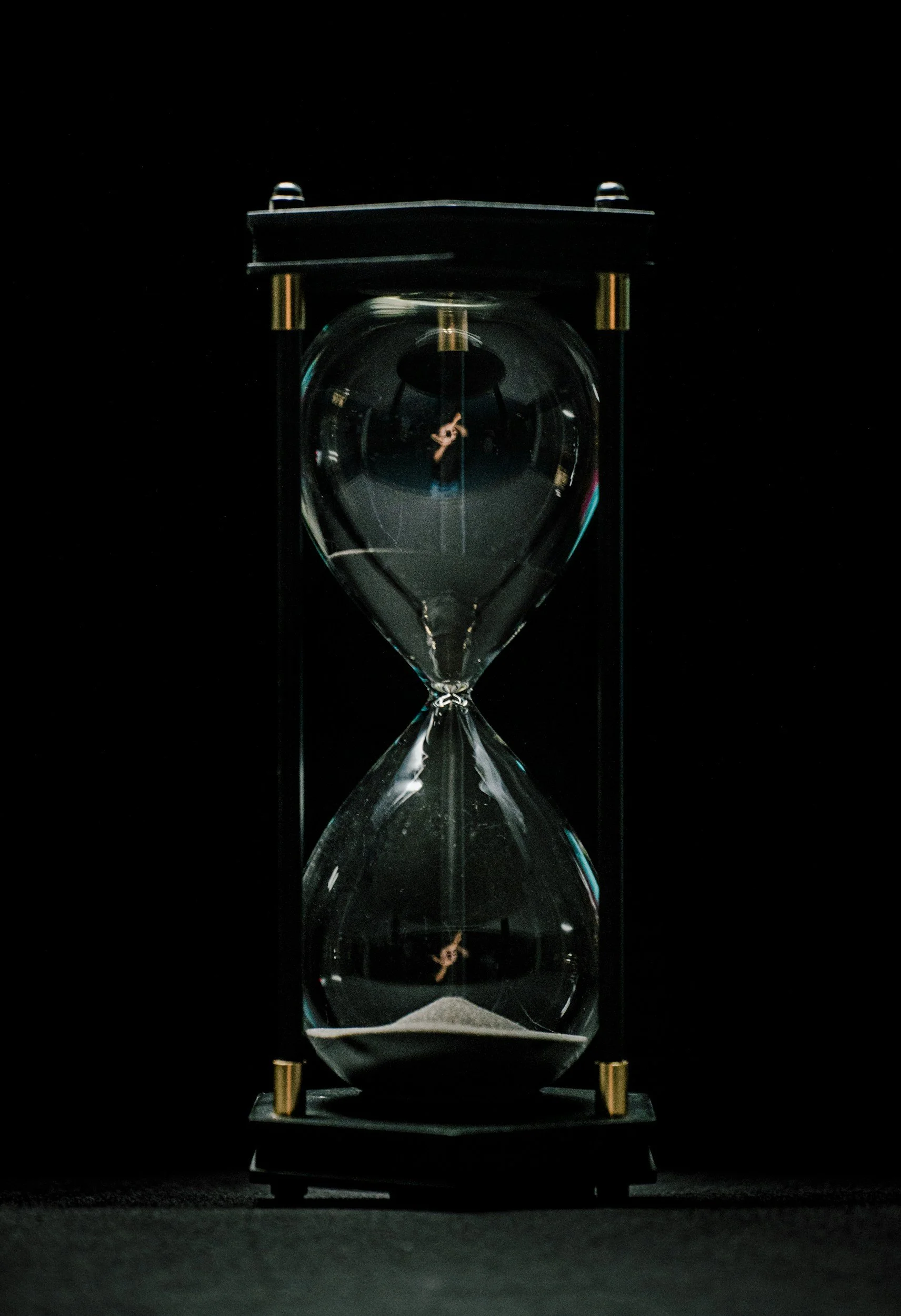 An hourglass with black frame and glass bulbs, filled with gray sand at the bottom, against a black background.