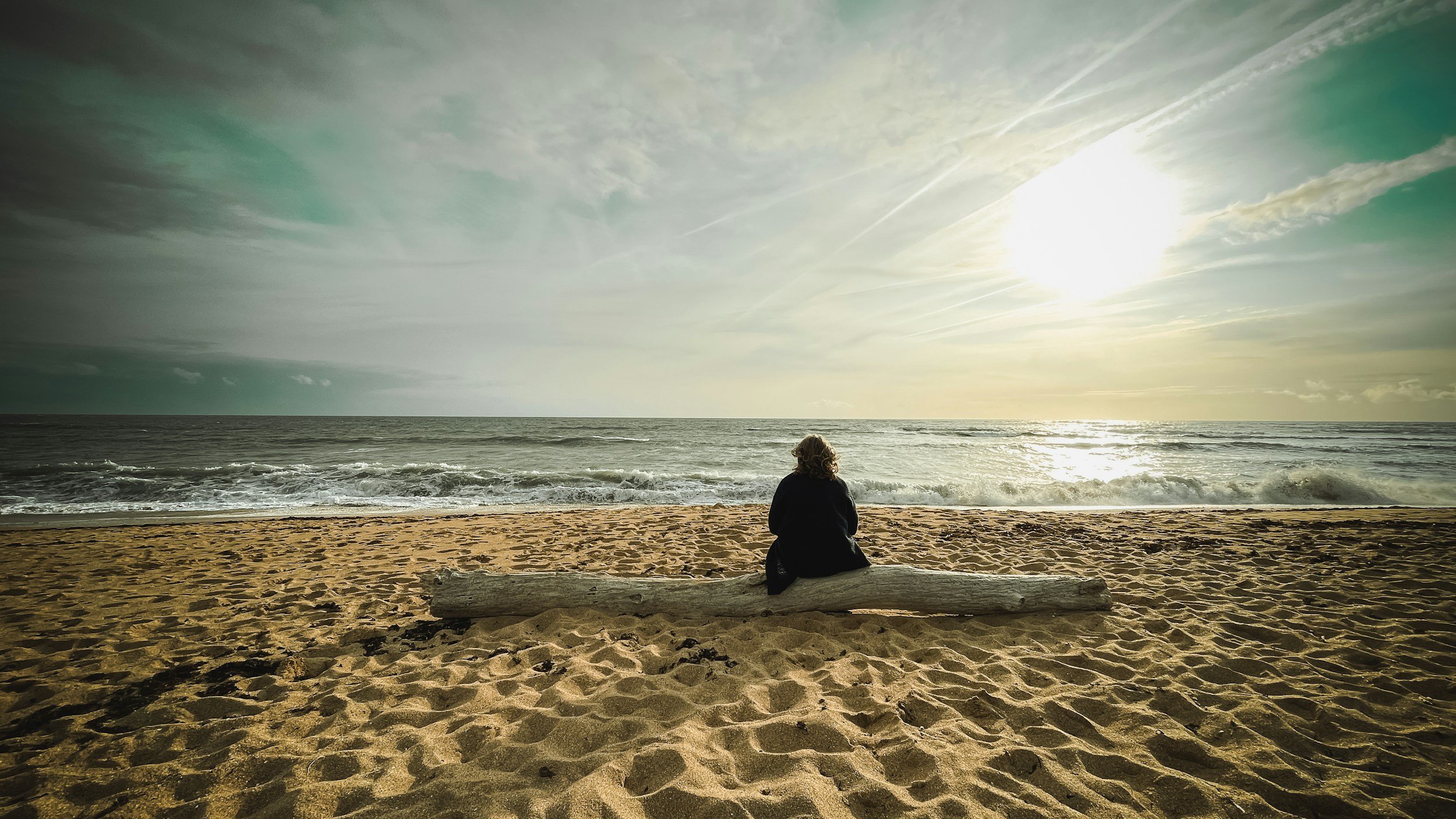 A person sitting on a driftwood log on a sandy beach, looking at the ocean with waves and a partly cloudy sky during sunset or sunrise.