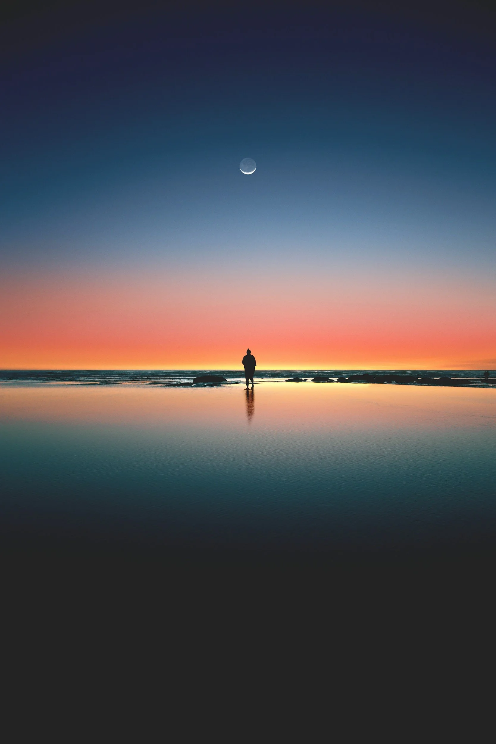 Silhouette of a person standing on the beach during sunset with a colorful sky, moon, and reflection on wet sand.