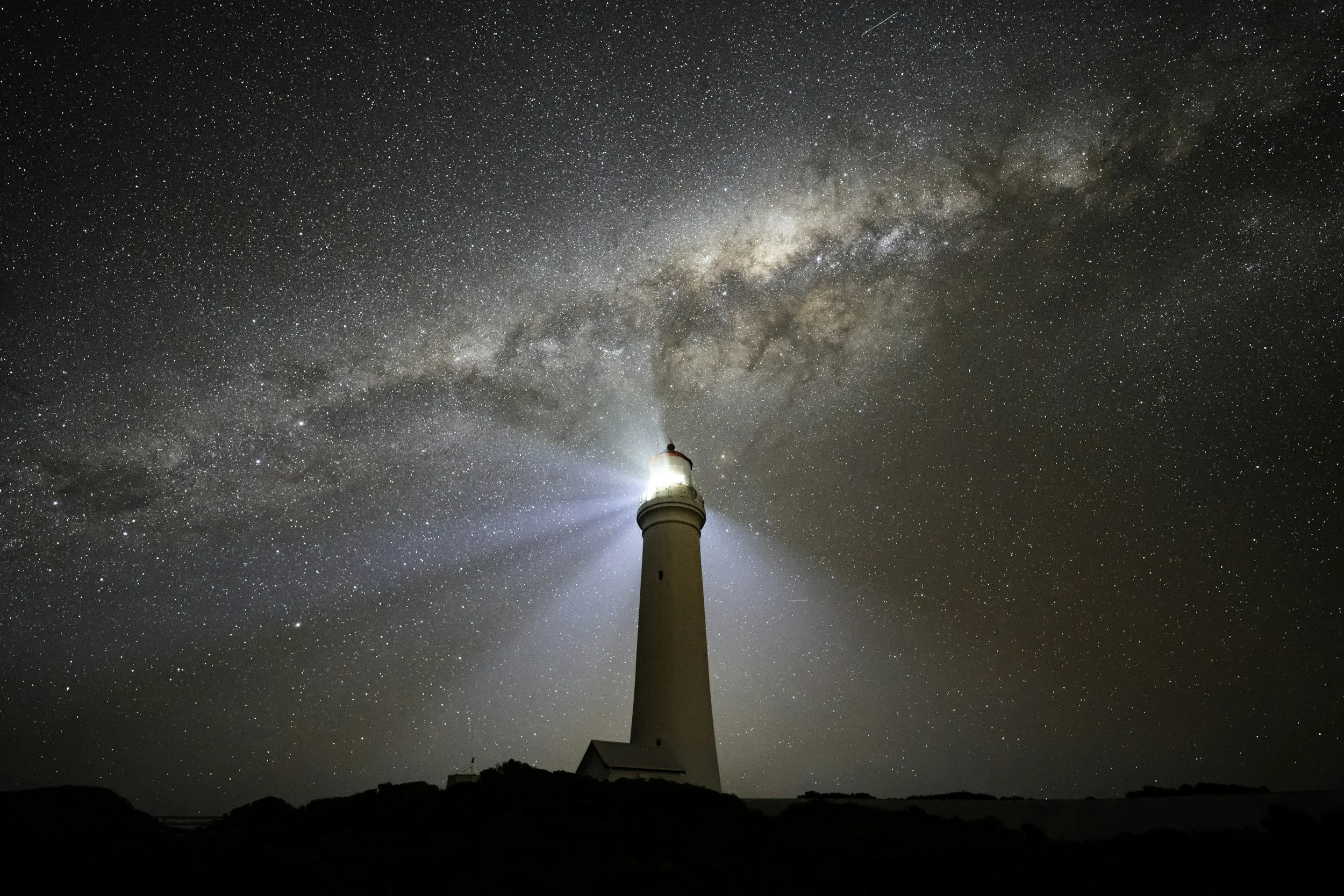 A lighthouse illuminated under a starry night sky with the Milky Way galaxy visible overhead.