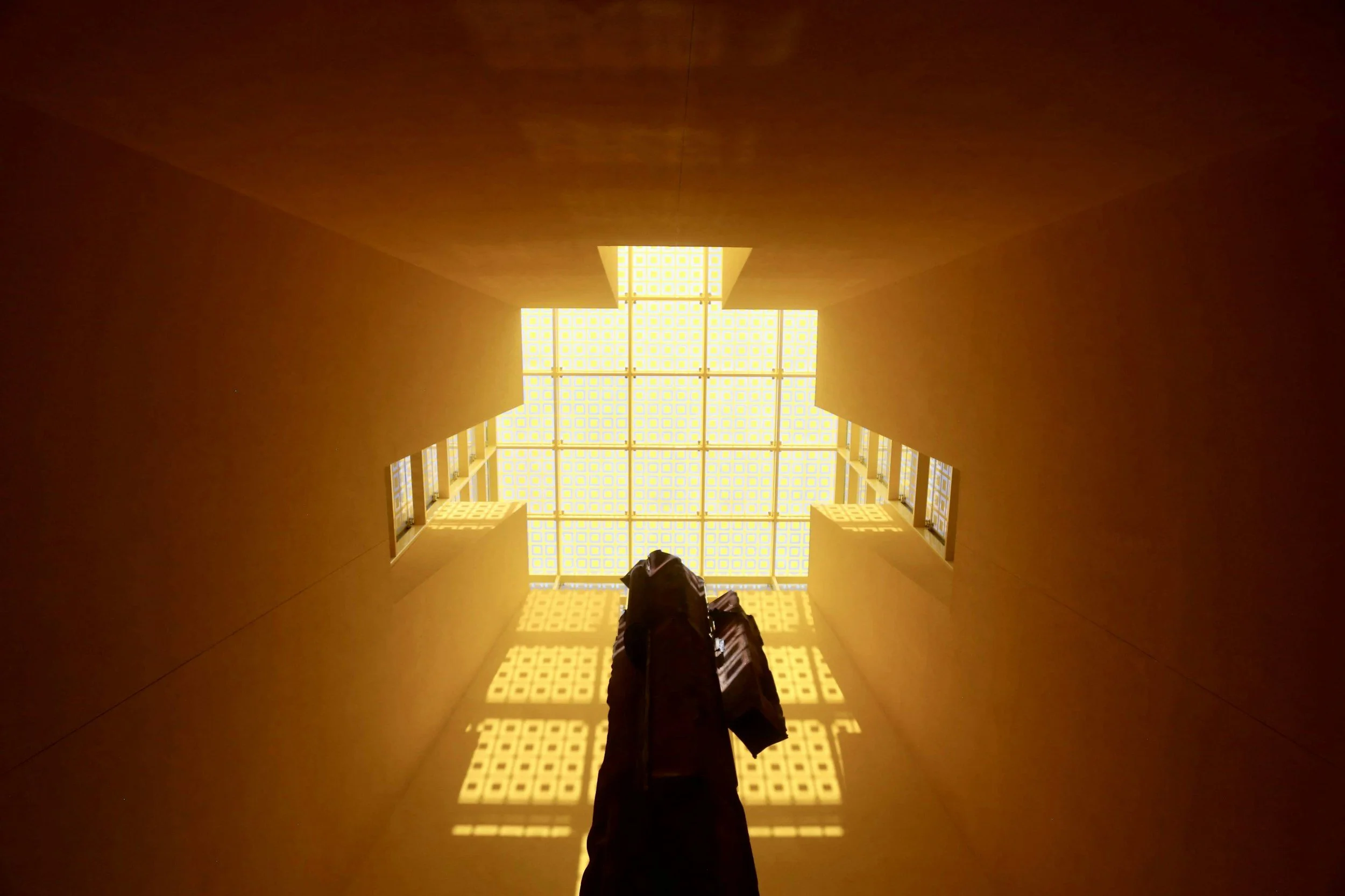View looking up at a bright yellow ceiling with square glass blocks, with some orange walls and a person in dark clothing and a bag standing underneath.