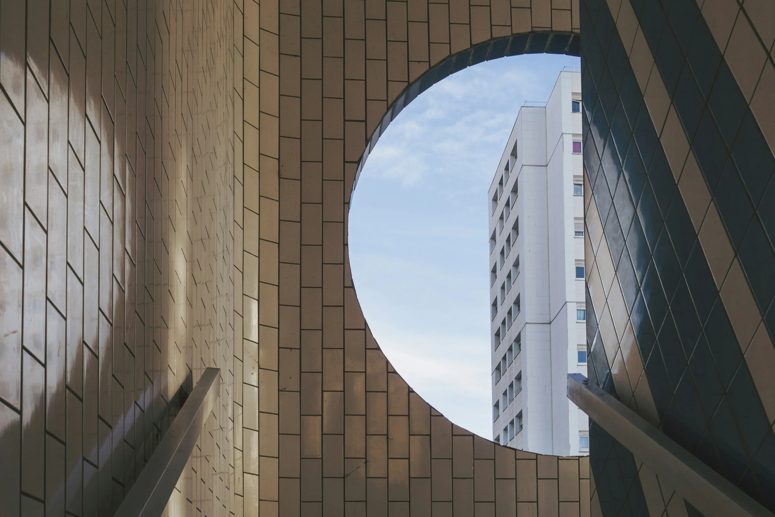 Looking up through a circular opening in a building with brick walls, revealing a neighboring white high-rise building with windows against a cloudy sky.