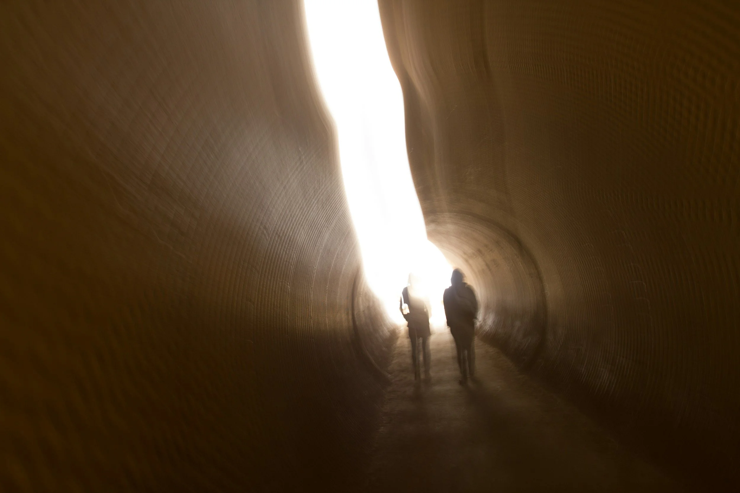 Two people walking through a brightly lit tunnel, viewed from inside, with the light creating silhouettes of the figures.
